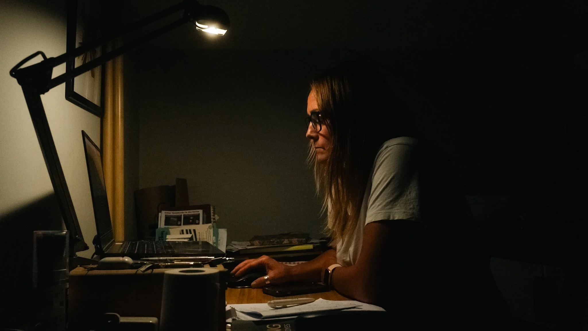 A woman working at her desk in a dimly lit room, focused on her computer screen.