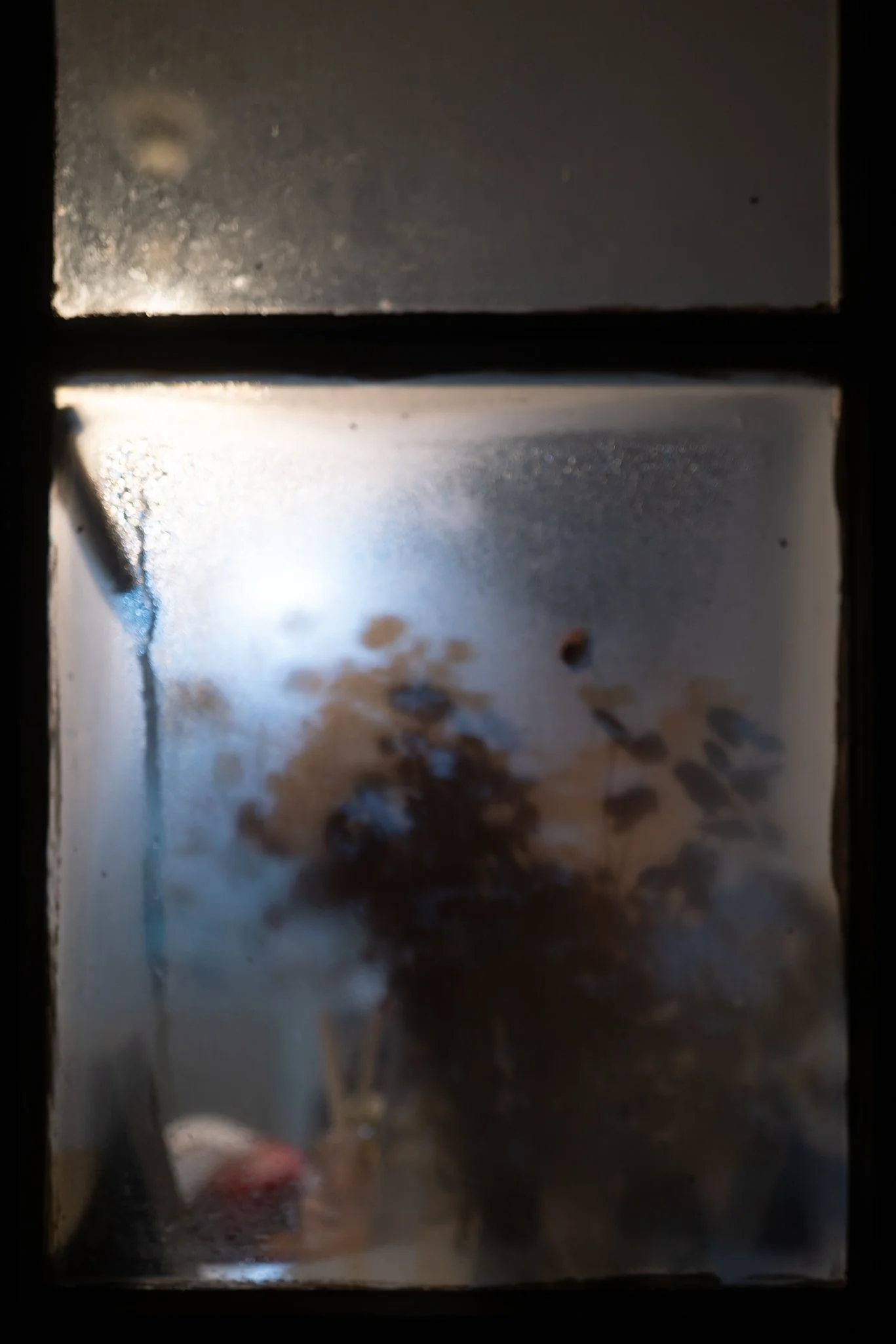 A blurry view through a window with condensation and smudges, showing a dark object possibly a figure, and some indistinct objects in the background.