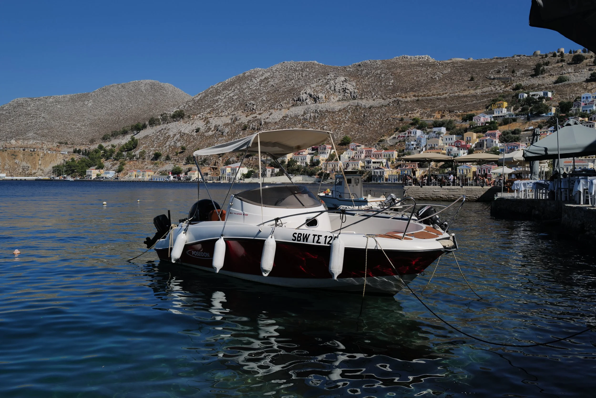 A small motorboat docked in a harbor with colorful houses on a hillside and rugged mountains in the background.