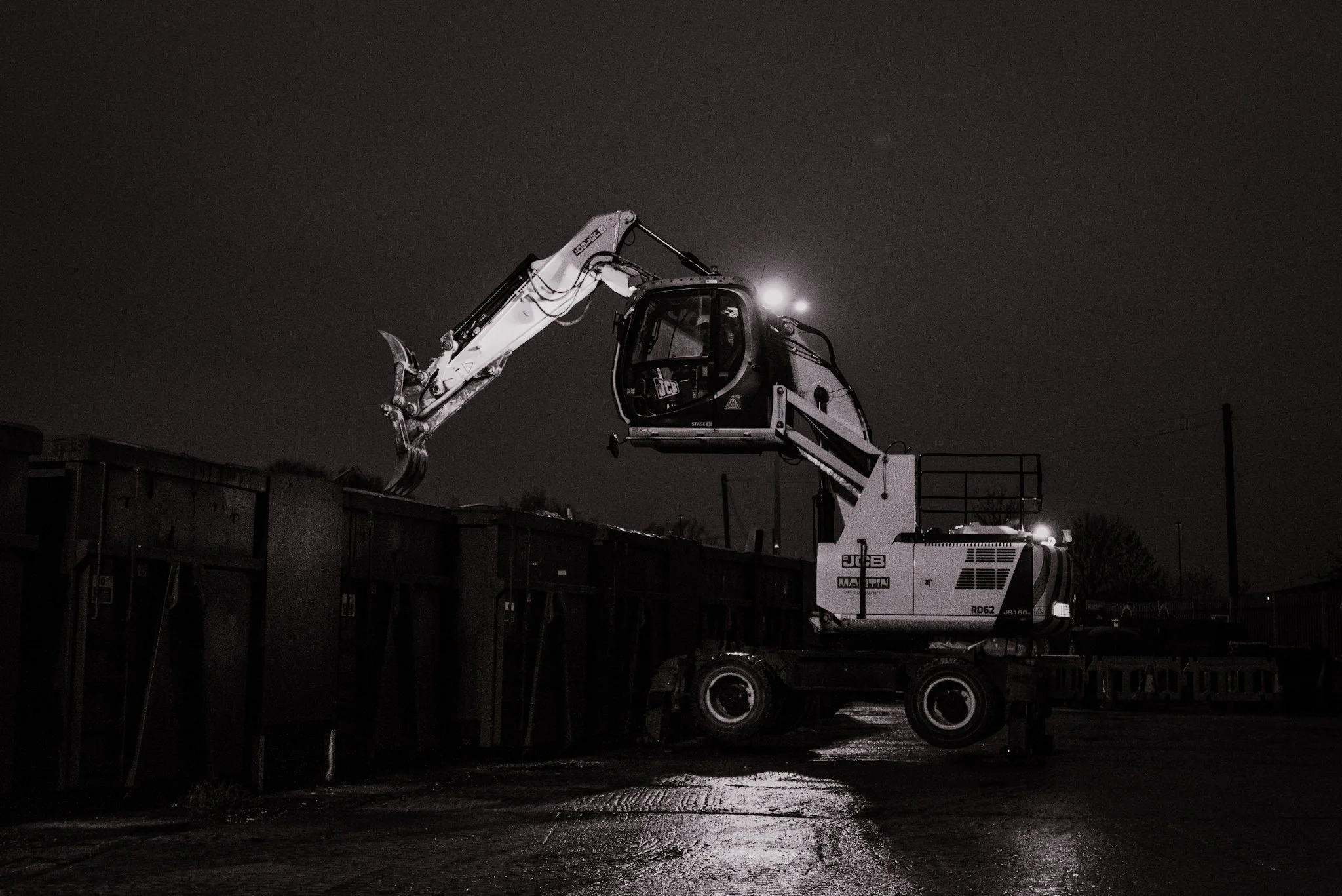 A large excavator at night, lifting debris off a tall container with its arm extended.