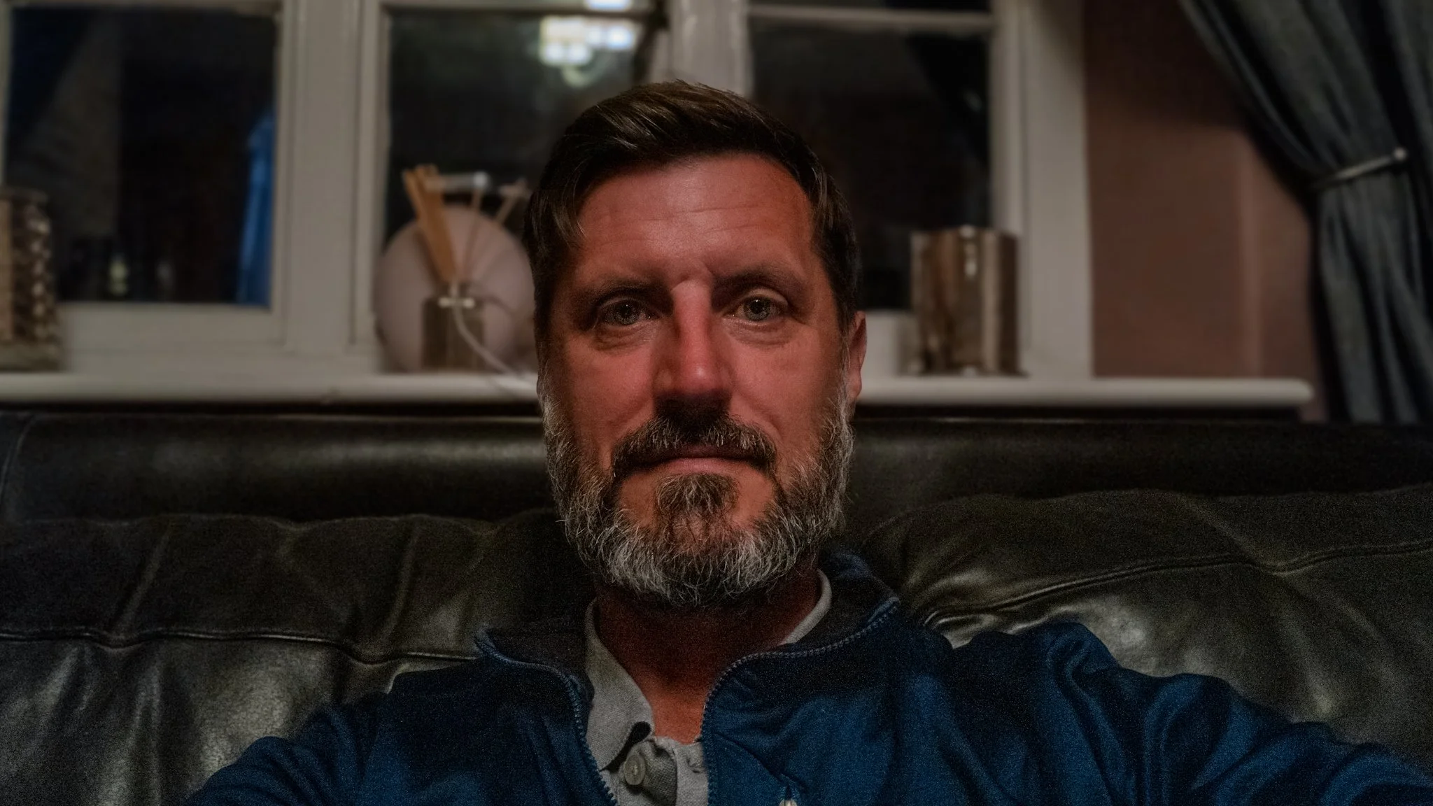 Close-up of a middle-aged man with a beard and mustache sitting on a black leather couch in a dimly lit room, looking at the camera.