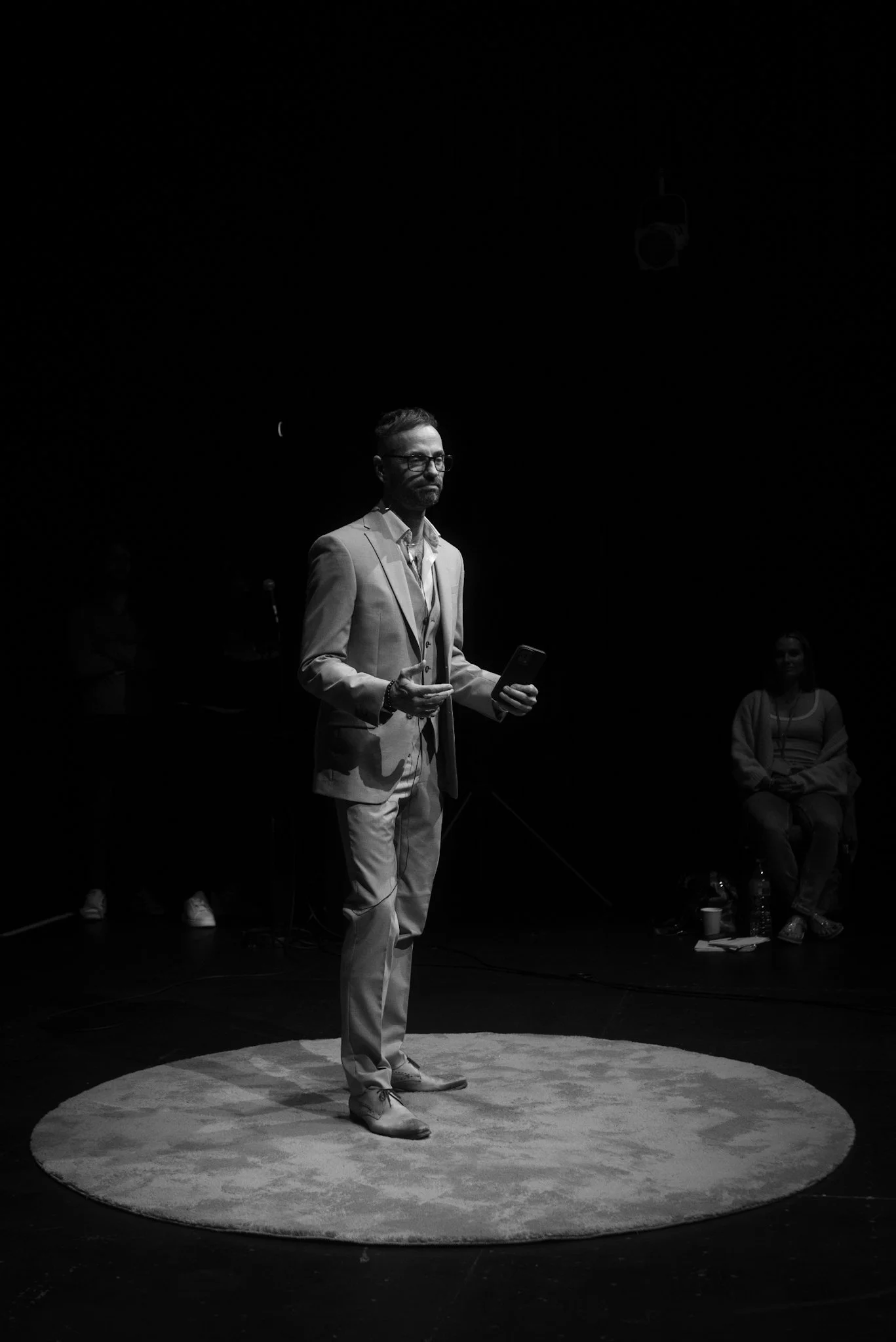 A man dressed in a suit holds a microphone and is standing on a circular rug on a dark stage, possibly giving a presentation or speech.