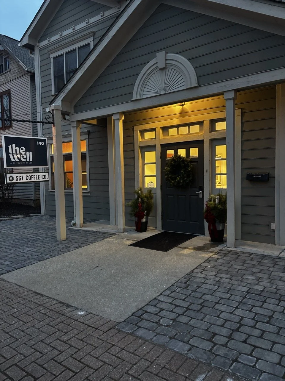 A house entrance illuminated with warm yellow lights, decorated with a wreath on the door, surrounded by potted plants, with a sign for 'the well' and 'SGT coffee co.' outside.