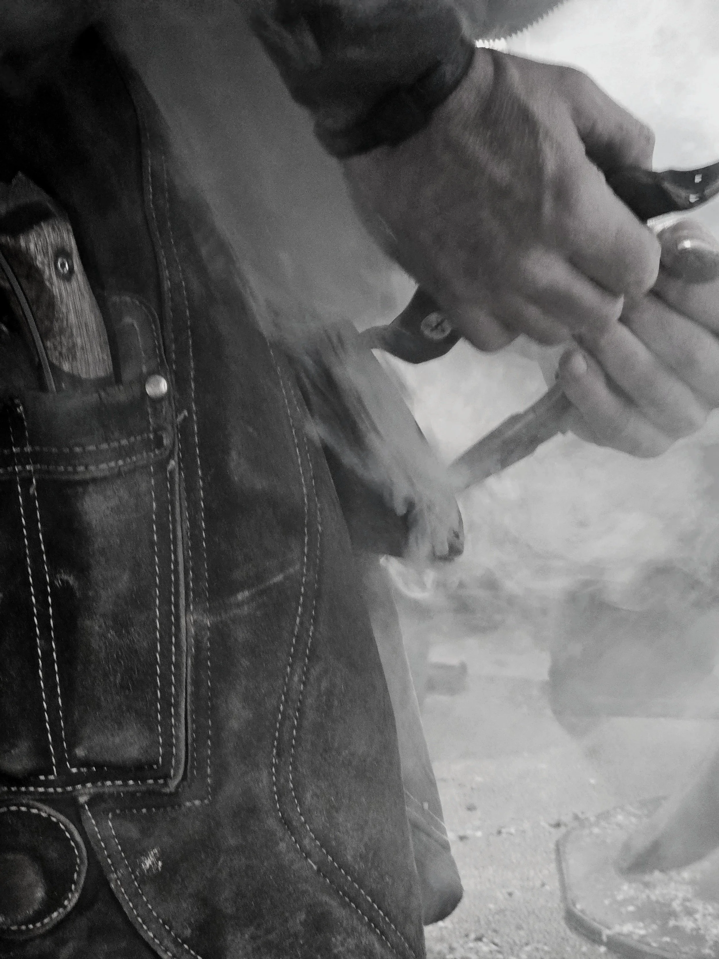 Black and white photo showing a person holding a handgun near their waist with their right hand, wearing jeans, and standing on a concrete surface.
