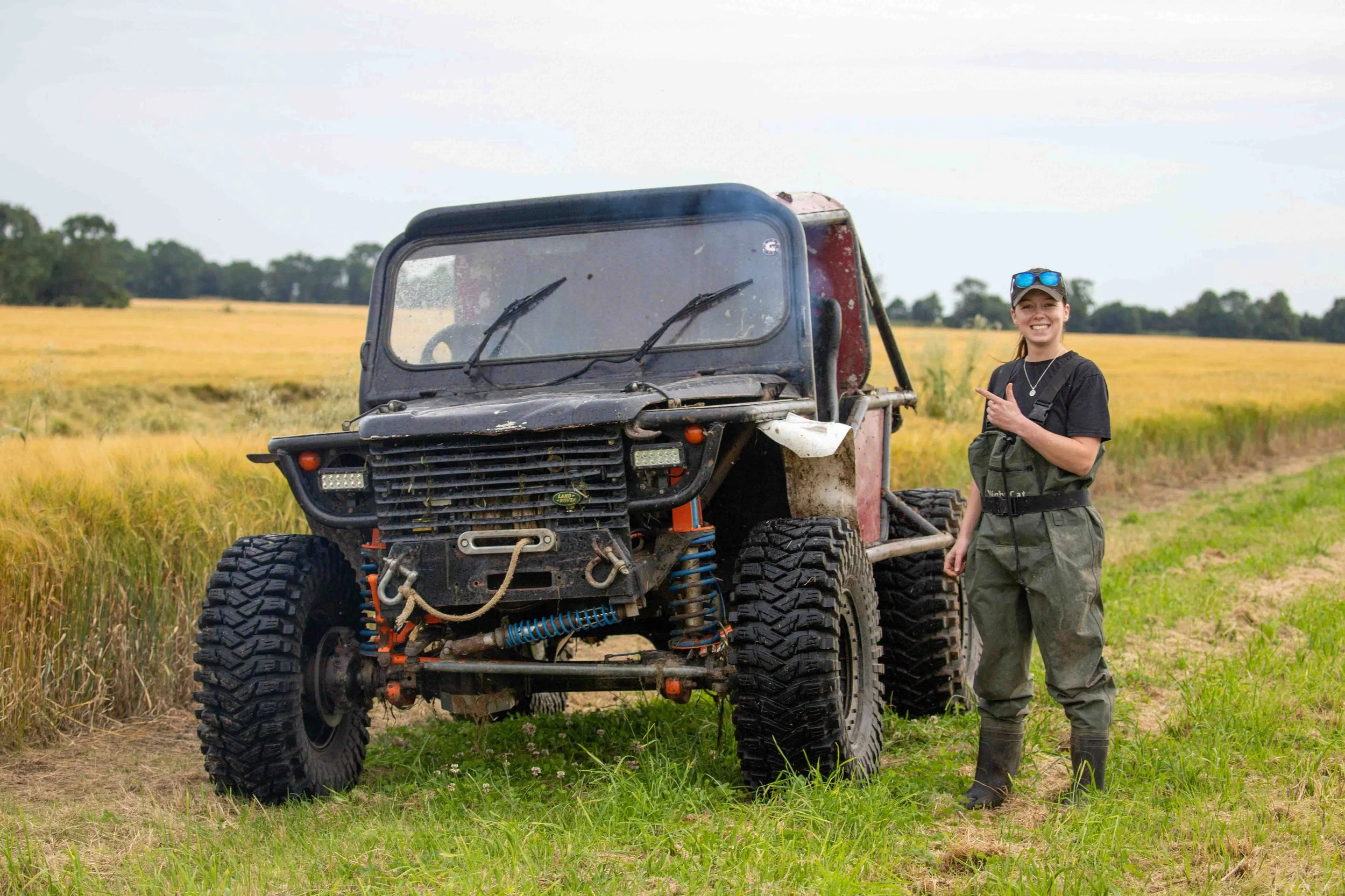 A woman in outdoor gear smiling and pointing at a damaged off-road utility vehicle in a grassy field with trees in the background.