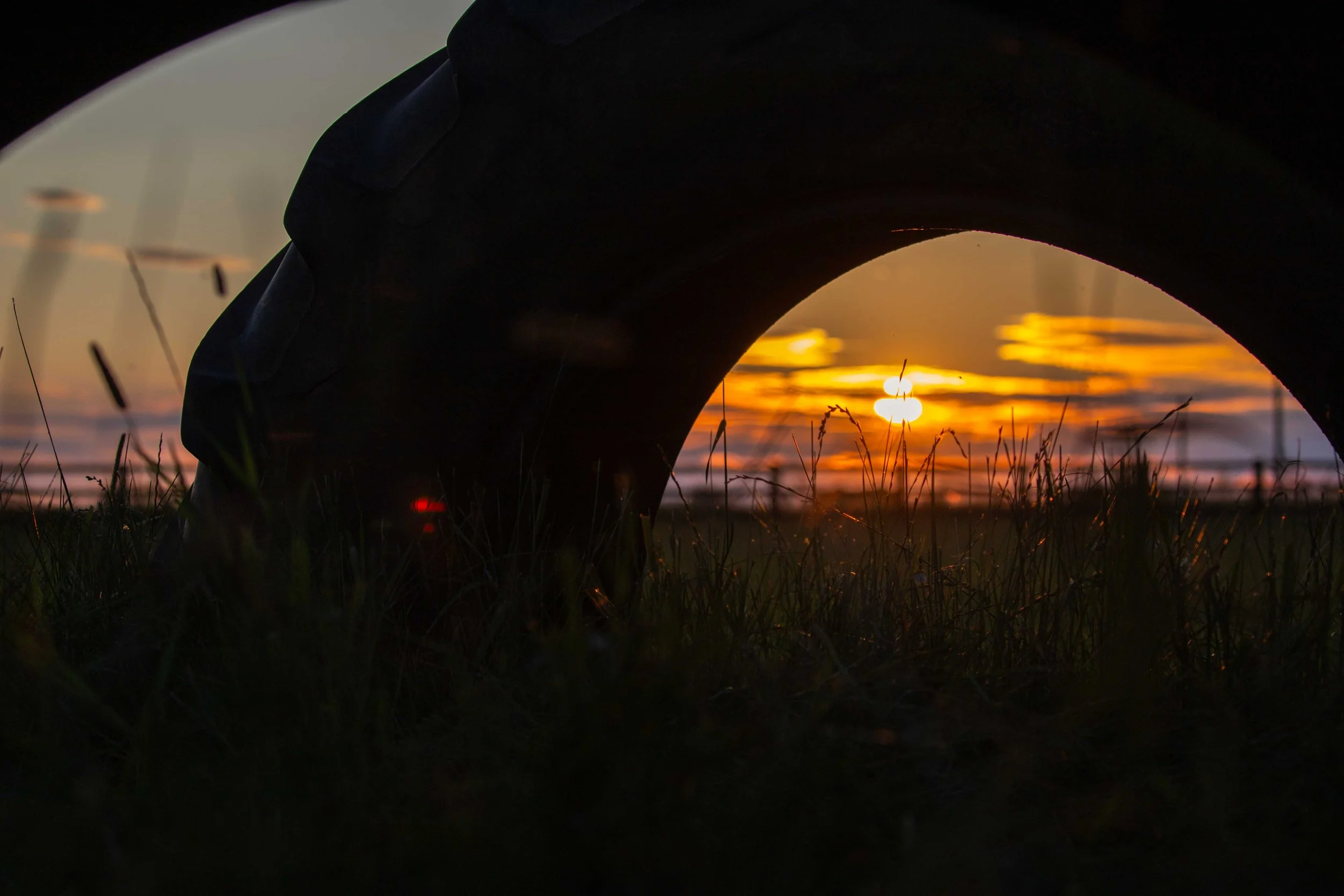 A close-up outdoor scene during sunset with the sun near the horizon, orange and yellow sky, and silhouettes of grass and a dark structure in the foreground.