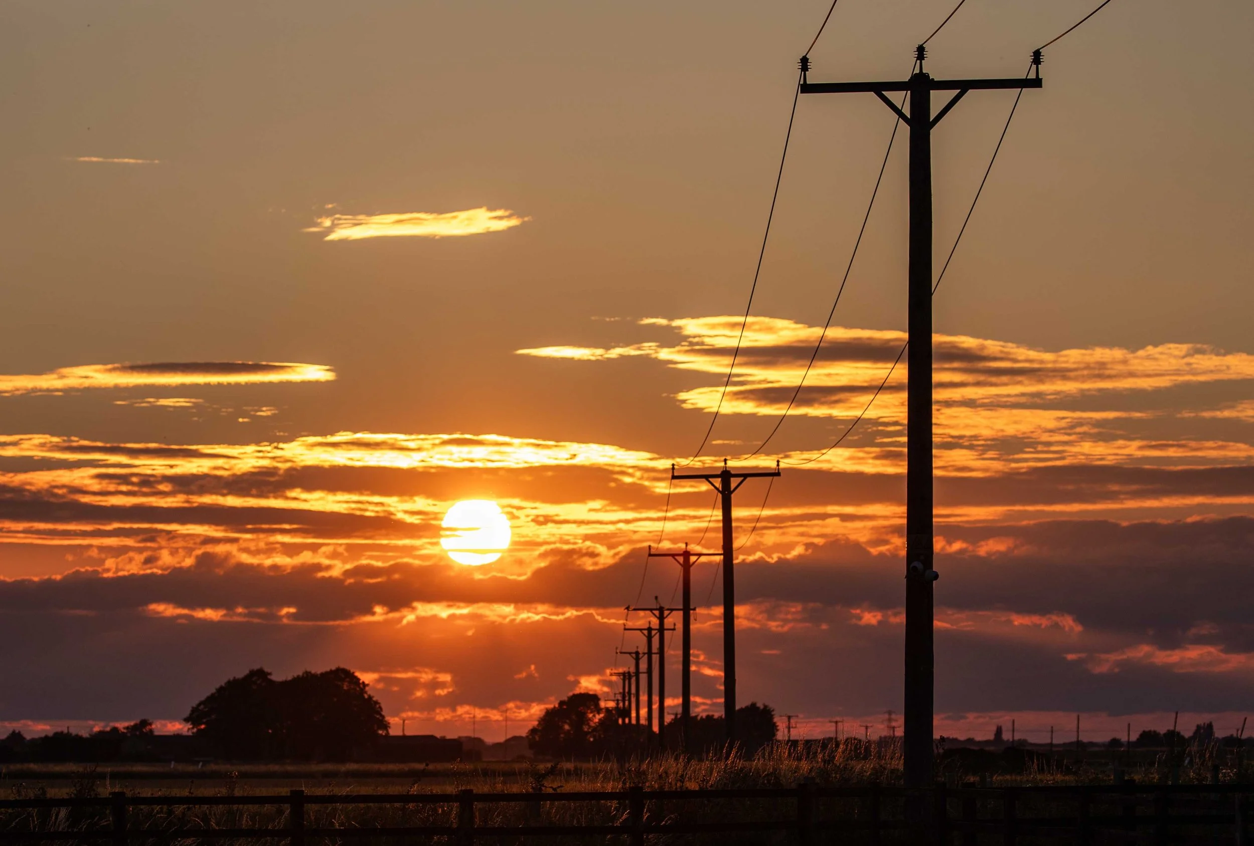 Sunset scene with power lines and utility poles against a sky with clouds illuminated by the setting sun.