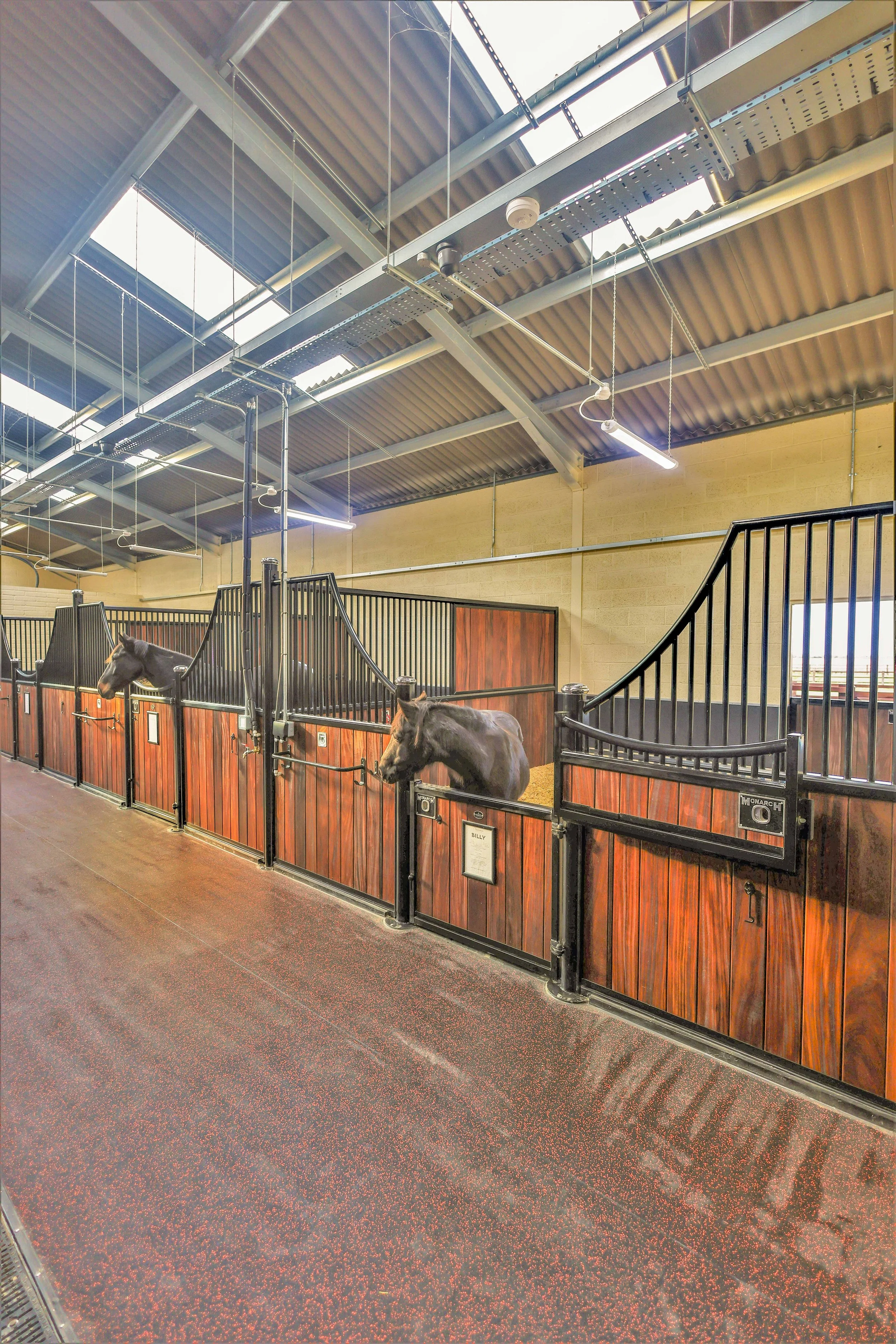 Interior of a stable with horses in individual stalls, tags with names and information on each stall, a brick wall and metal railings, and a clean rubberized floor with red speckling.