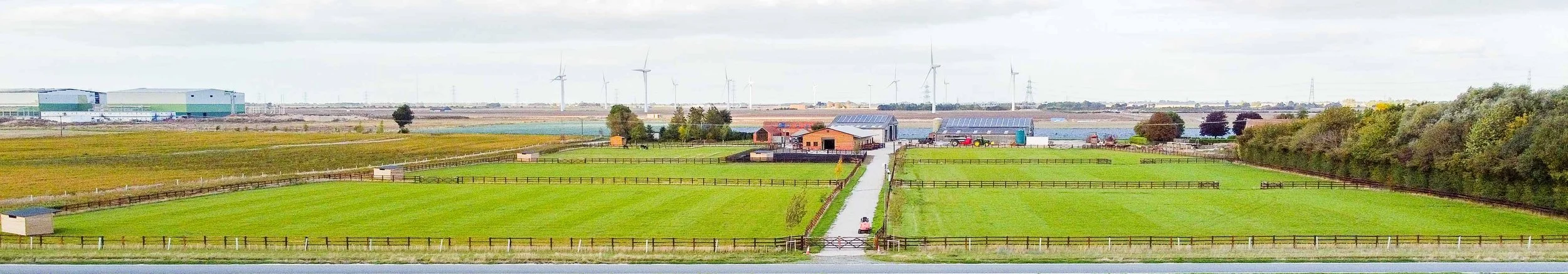 A rural landscape with green pastures, farm buildings, and wind turbines in the distance under a cloudy sky.
