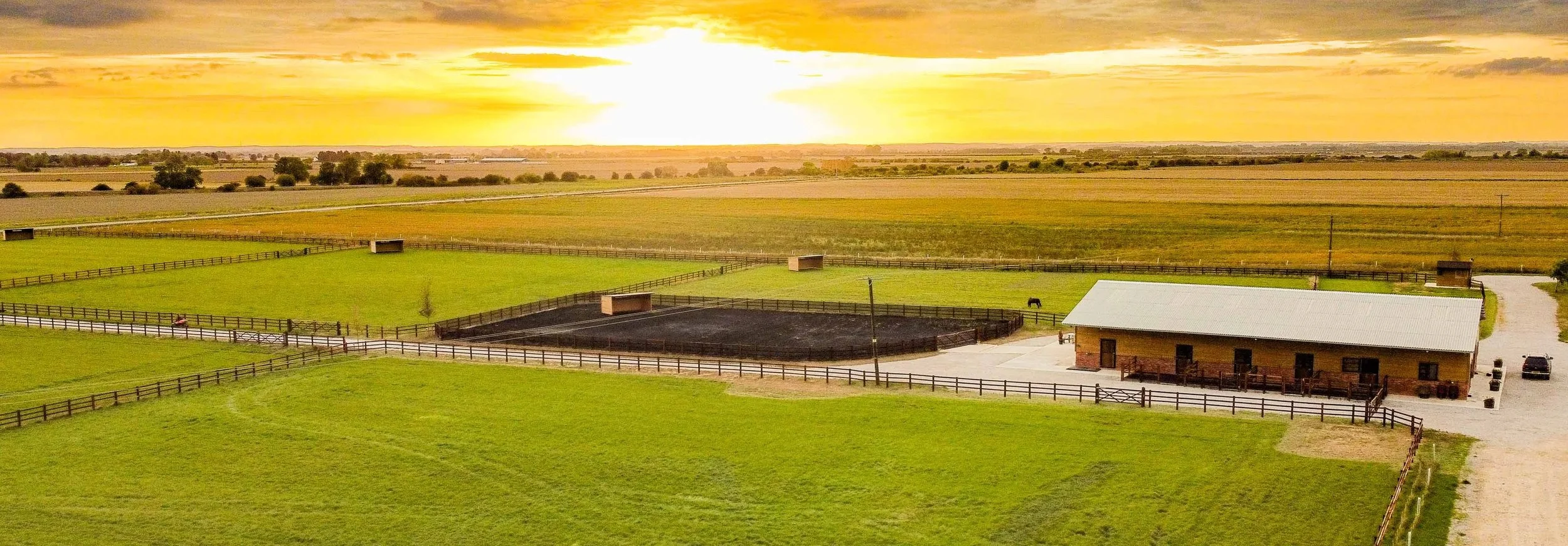 A rural farm with a green pasture, a small barn, a fenced riding area, and a building, all under a dramatic sunset sky.