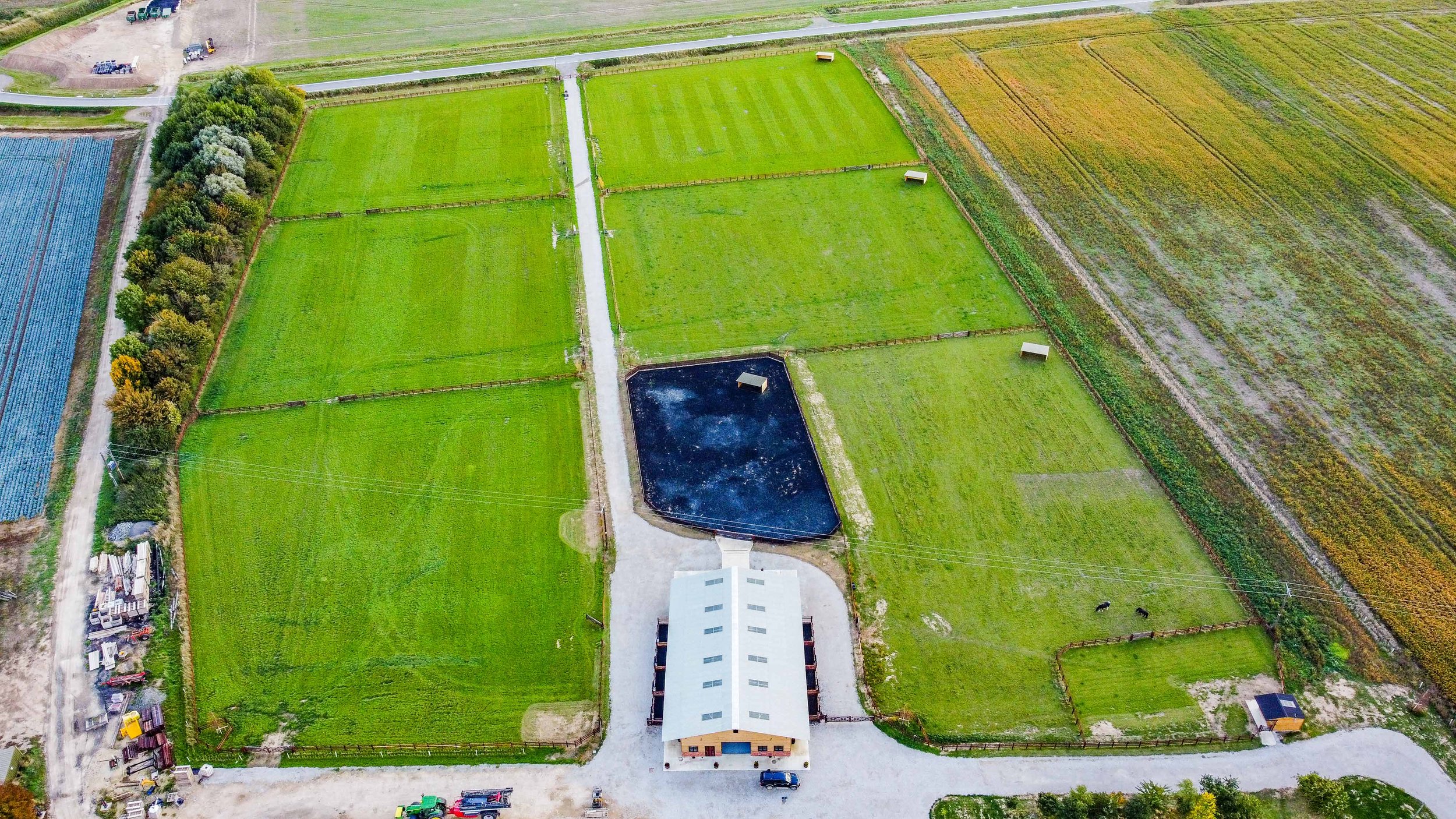 Aerial view of a farm with green fields, a black water pond, a large white barn, and surrounding trees and dirt roads.