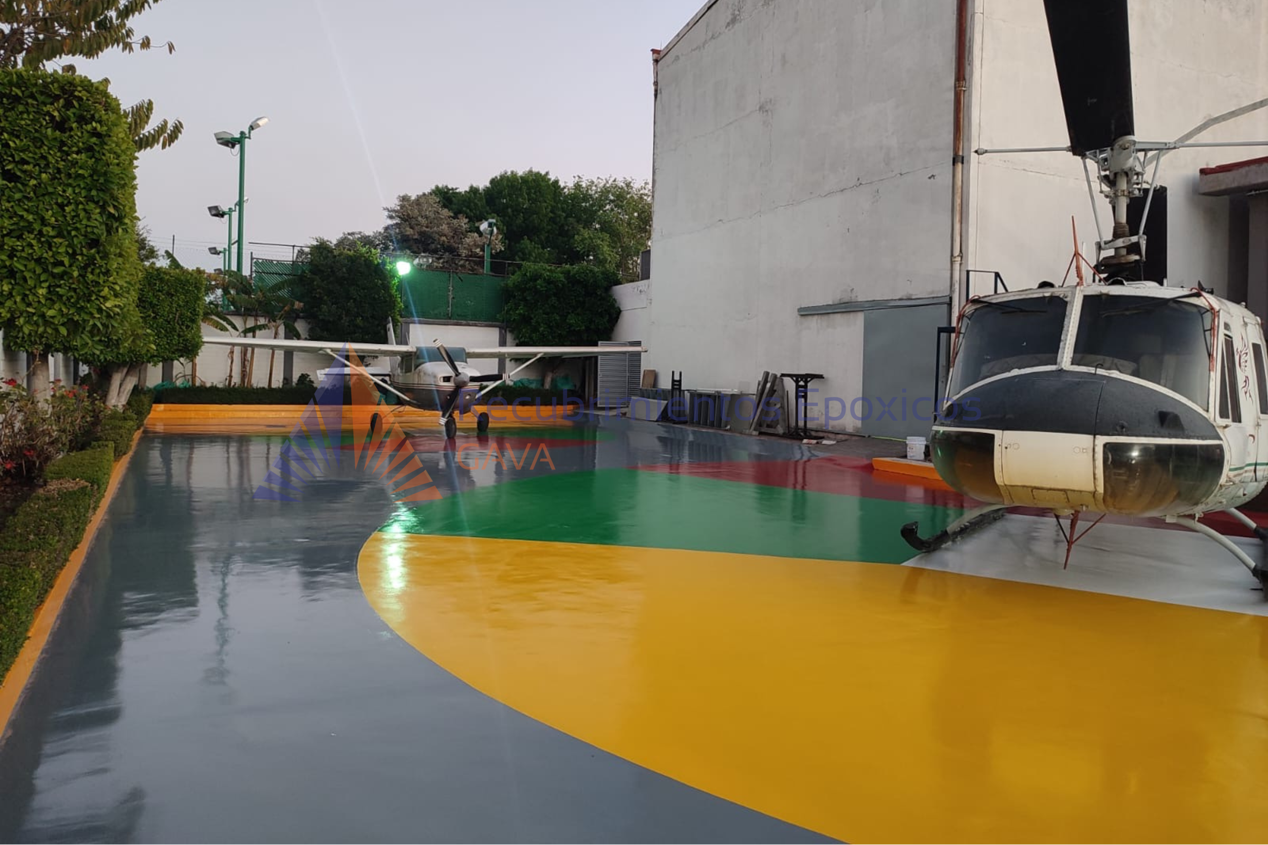 Helicóptero sobre pista de aeropuerto, con colores amarillo, verde y gris, cercada por árboles y edificaciones.