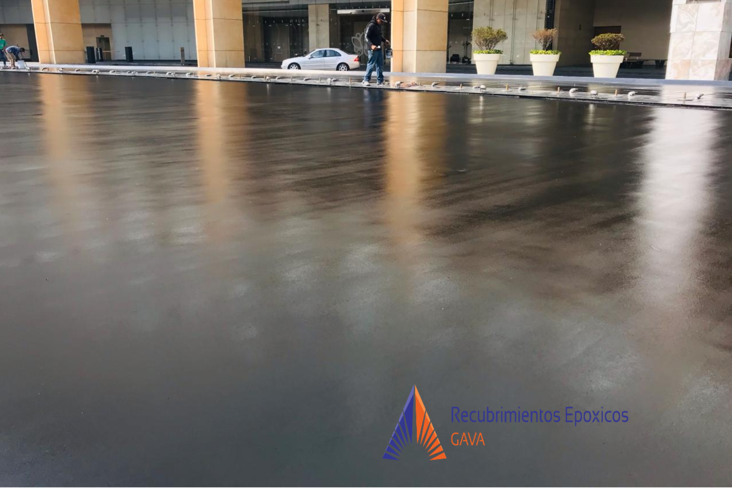 Proceso de recubrimiento epóxico en una superficie de piso en un área industrial o comercial. Se observa una capa de pintura de color negro brillante en el suelo, con dos personas en el fondo y plantas decorativas en macetas. En la parte inferior der