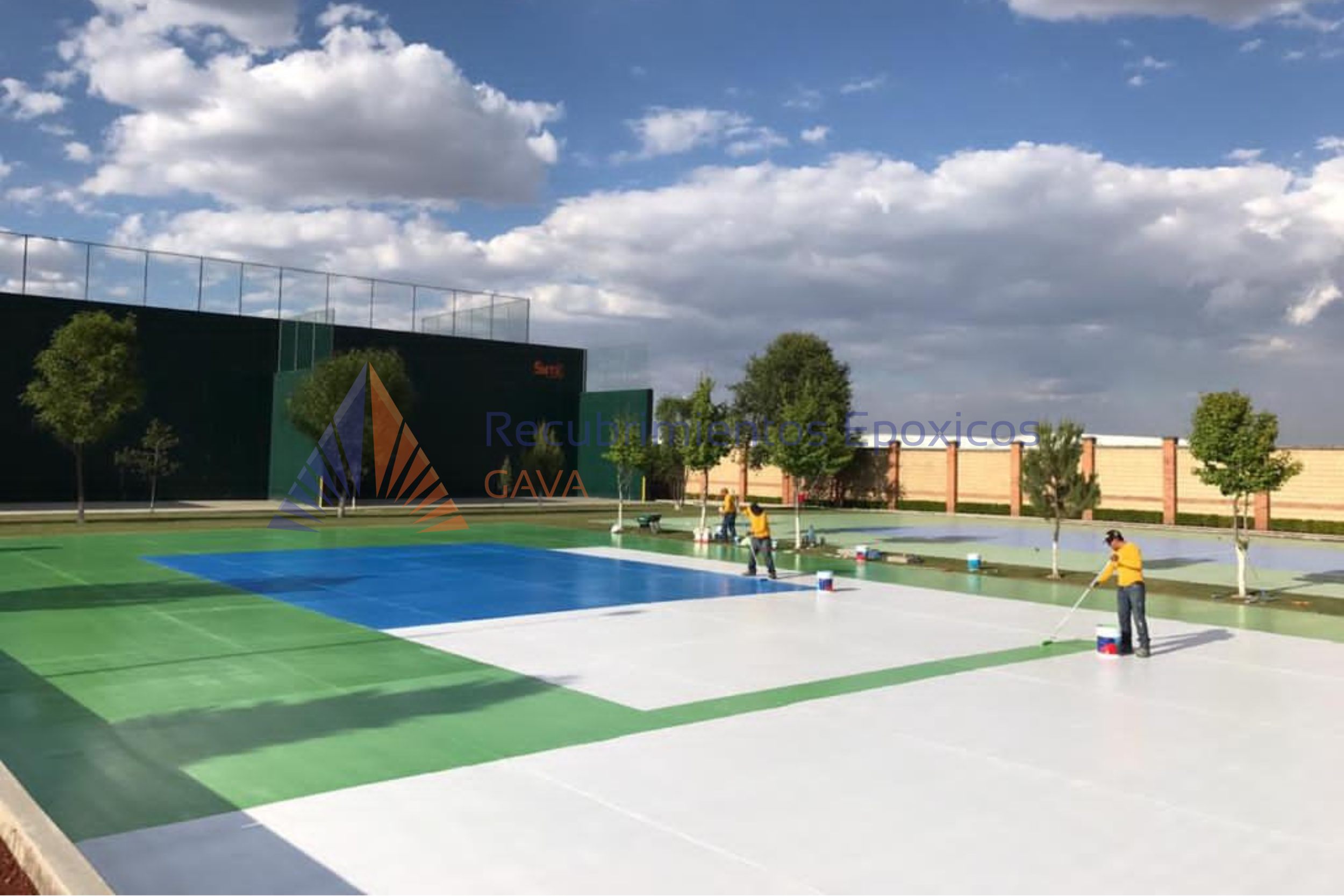 Personas pintando y limpiando canchas de tenis al aire libre en un día soleado con cielo nublado, árboles y estructuras deportivas en el fondo.