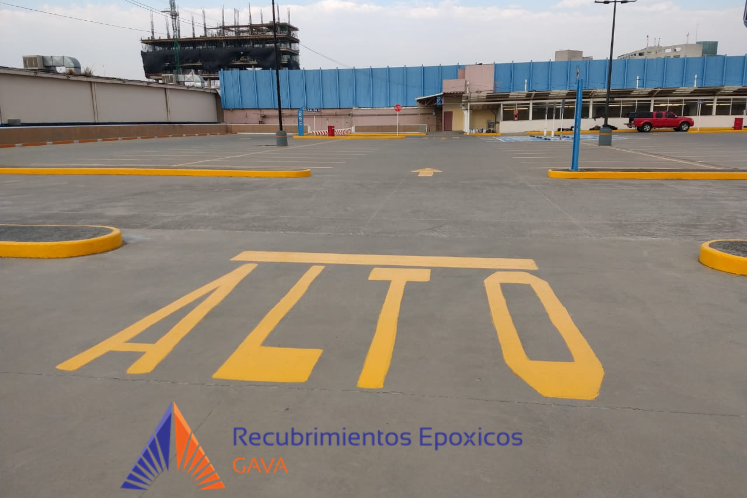 Estacionamiento vacío con señalización en amarillo y marcas de flechas en el suelo. Fondo con edificio con paredes de color beige y estructura de metal azul. Texto en la parte inferior que dice 'Recubrimientos Epóxicos GAVA' con un logo reluciente de
