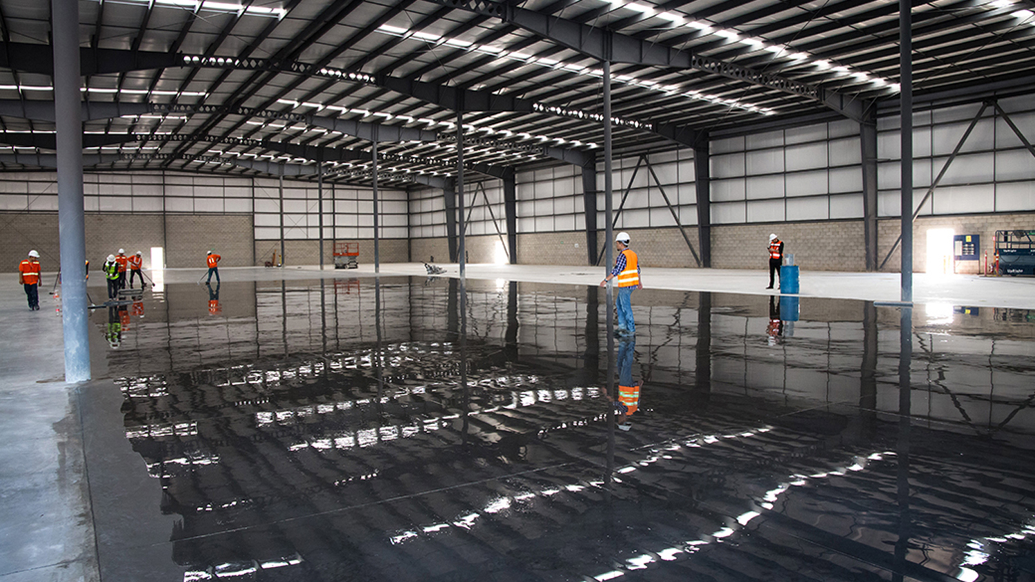 Personal de trabajo aplicando concreto en un galpón grande con estructura metálica y techos altos.