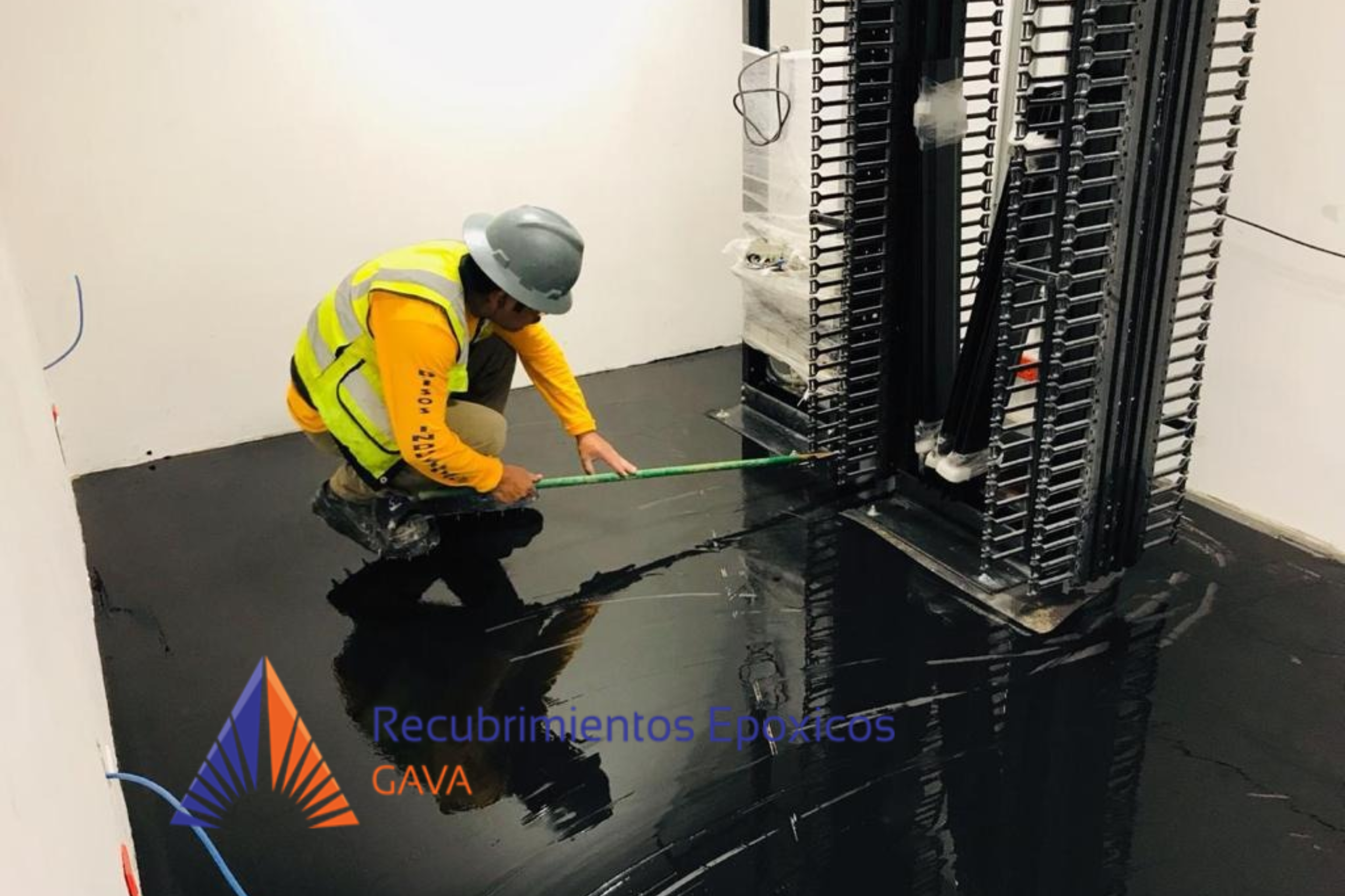 Un trabajador con casco y chaleco de seguridad aplicando recubrimiento epóxico negro en el suelo, junto a un panel de estructura metálica