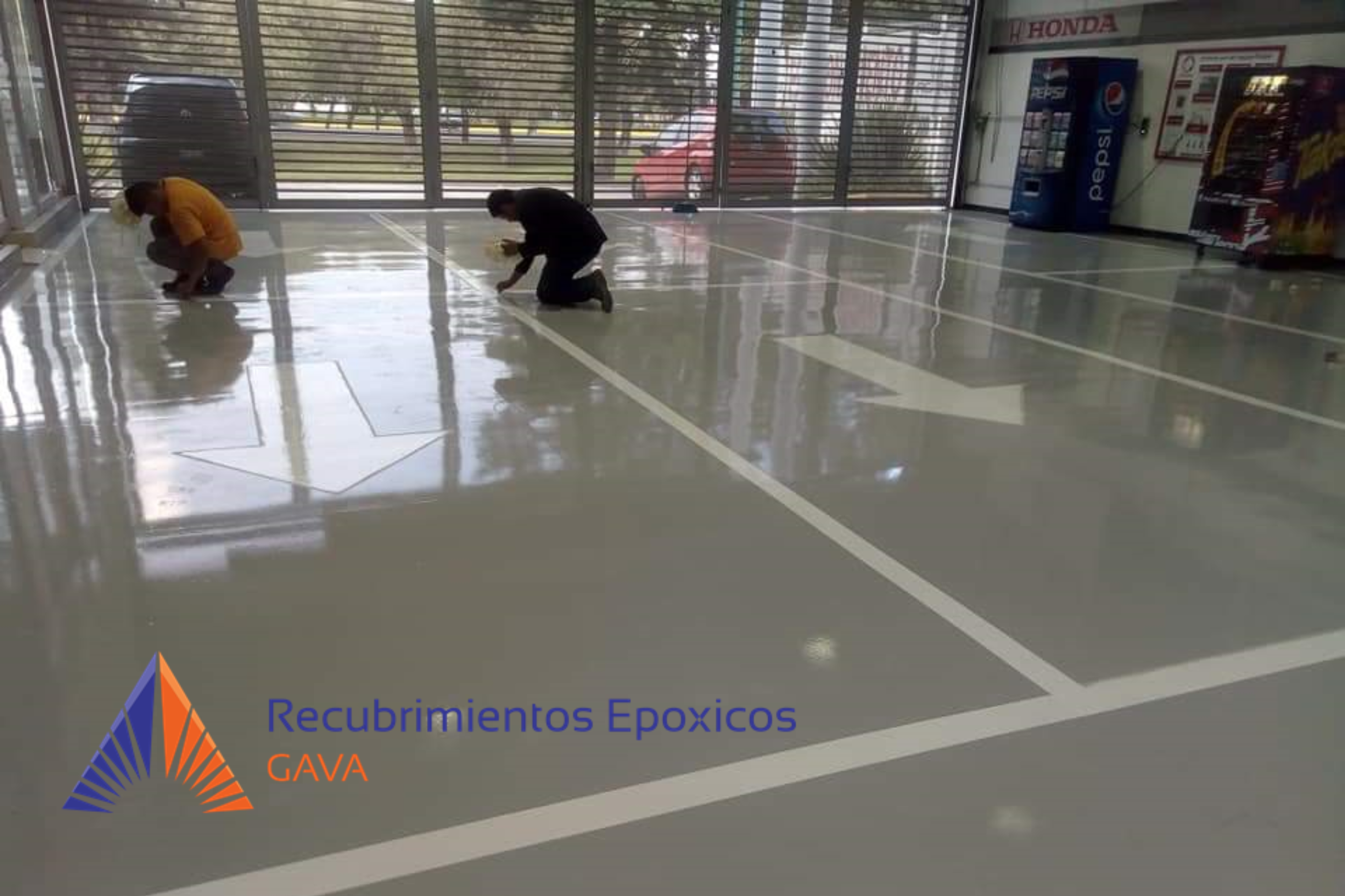 Dos personas trabajando en la preparación de recubrimientos epóxicos en un espacio cerrado con paredes de vidrio y puertas de colos, junto a máquinas expendedoras y un cartel con texto.