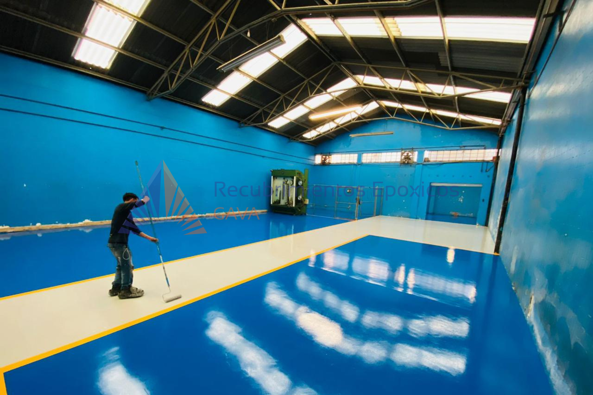 Interior de una cancha deportiva que está siendo pintada, con paredes azules y un techo a dos aguas con iluminación fluorescente. Hay una persona pintando en el piso con un rodillo.