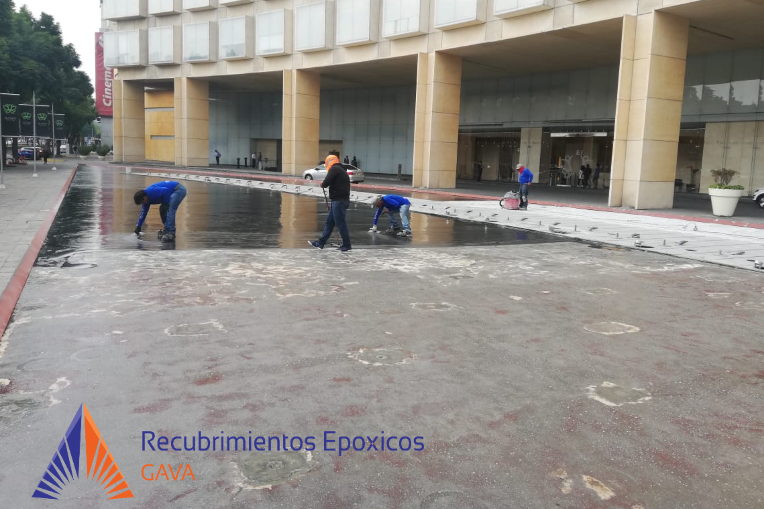 Personal de construcción aplicando recubrimiento epóxico en el piso de una calle frente a un edificio.