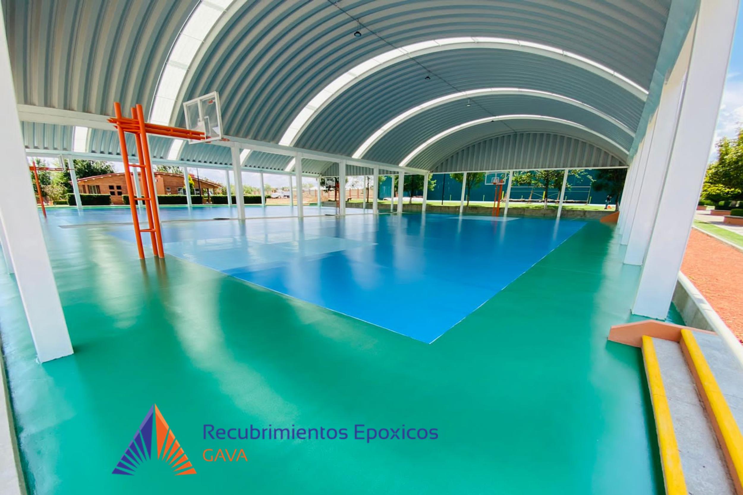 Pista de baloncesto cubierta con techo curvo, piso de color verde y azul, con arcos y aros. Hay vegetación y edificios en el fondo.