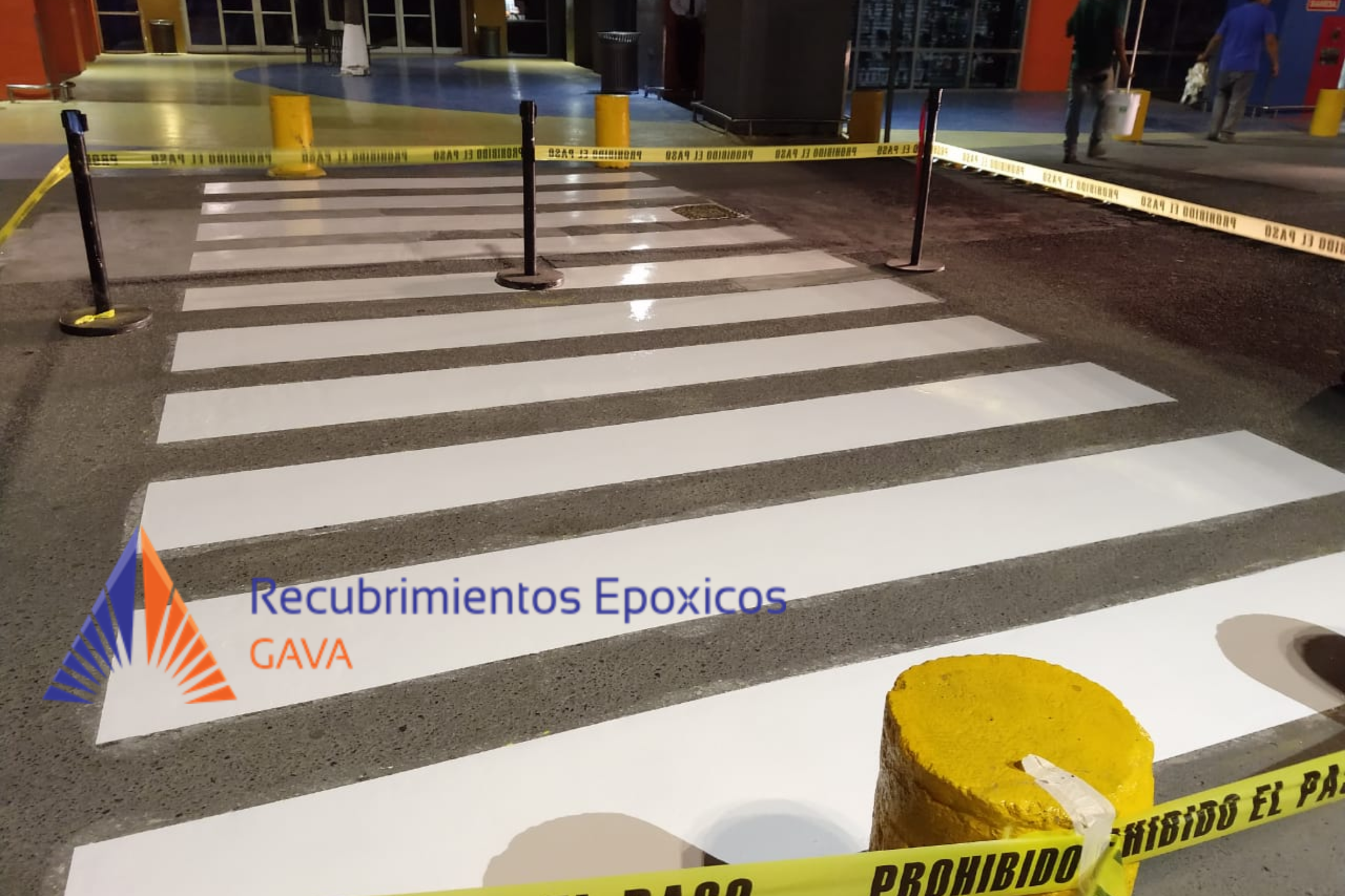 Corte de acceso peatonal con cinta de advertencia amarilla y postes, en la entrada de un edificio, con señalización y piso de mosaico colorida y un aviso de recubrimientos epóxicos en la esquina.