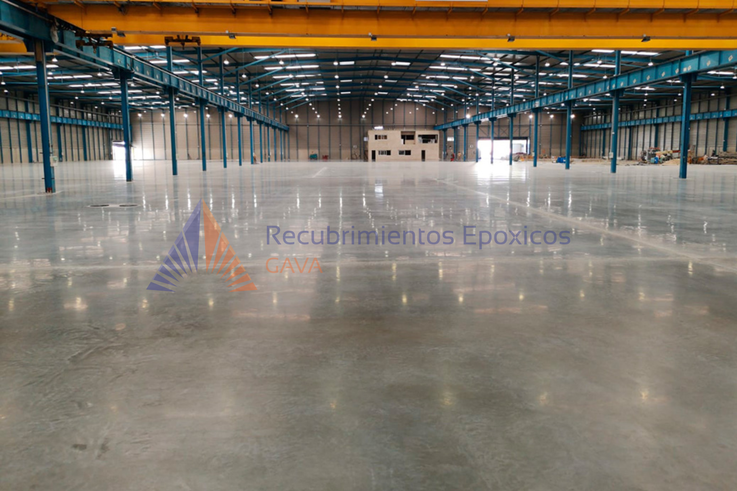 Interior de un almacén con estructura metálica azul y suelo de cemento pulido, con una oficina pequeña de madera al fondo y equipo en los lados. Inscripción en la parte inferior que dice "Recubrimientos Epóxicos GAVA".