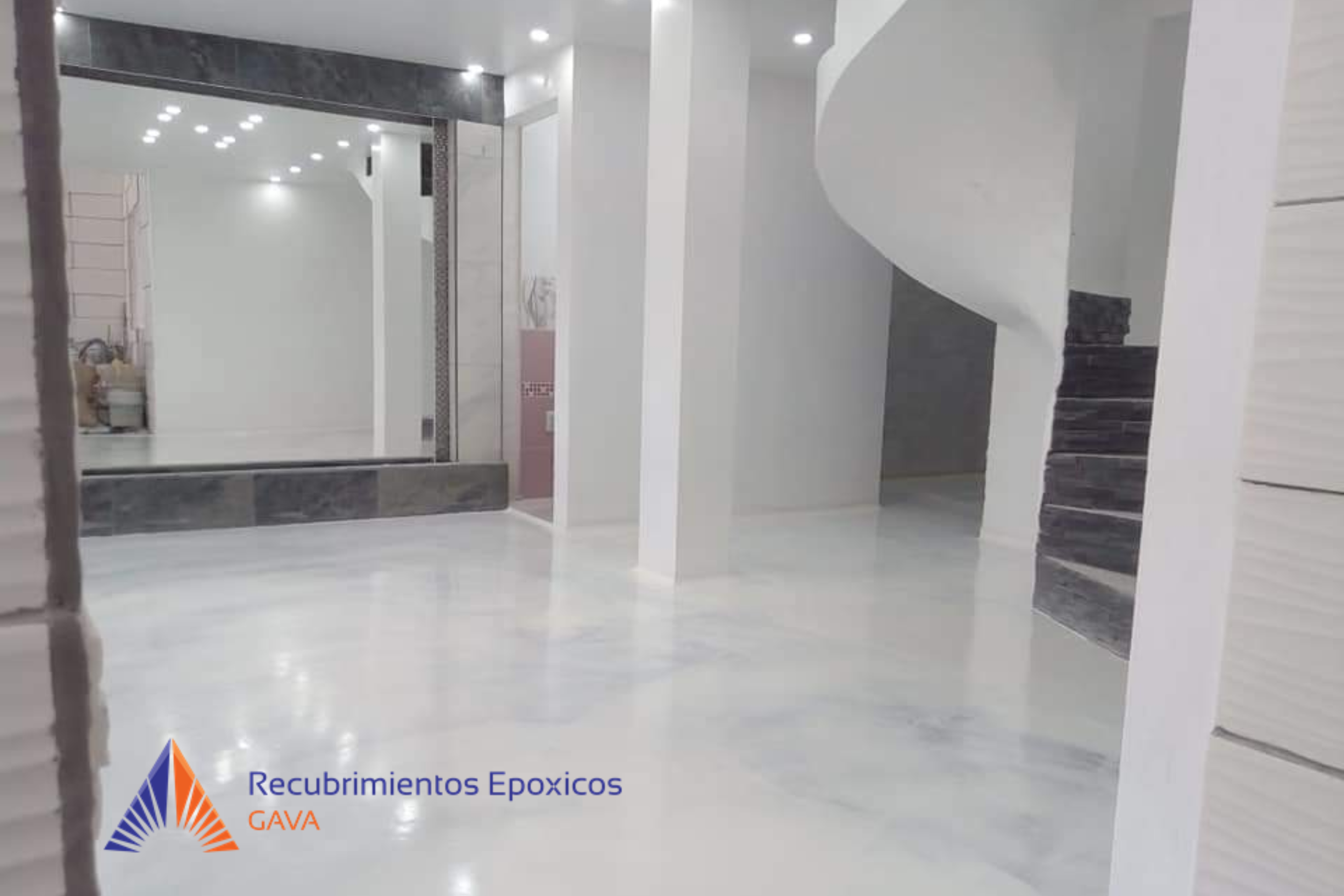 Interior de un edificio con paredes blancas y pisos de mármol claro, mostrando un espejo grande, iluminación moderna en el techo y una escalera de piedra negra. En la esquina inferior izquierda, hay un logotipo y texto de la empresa. 