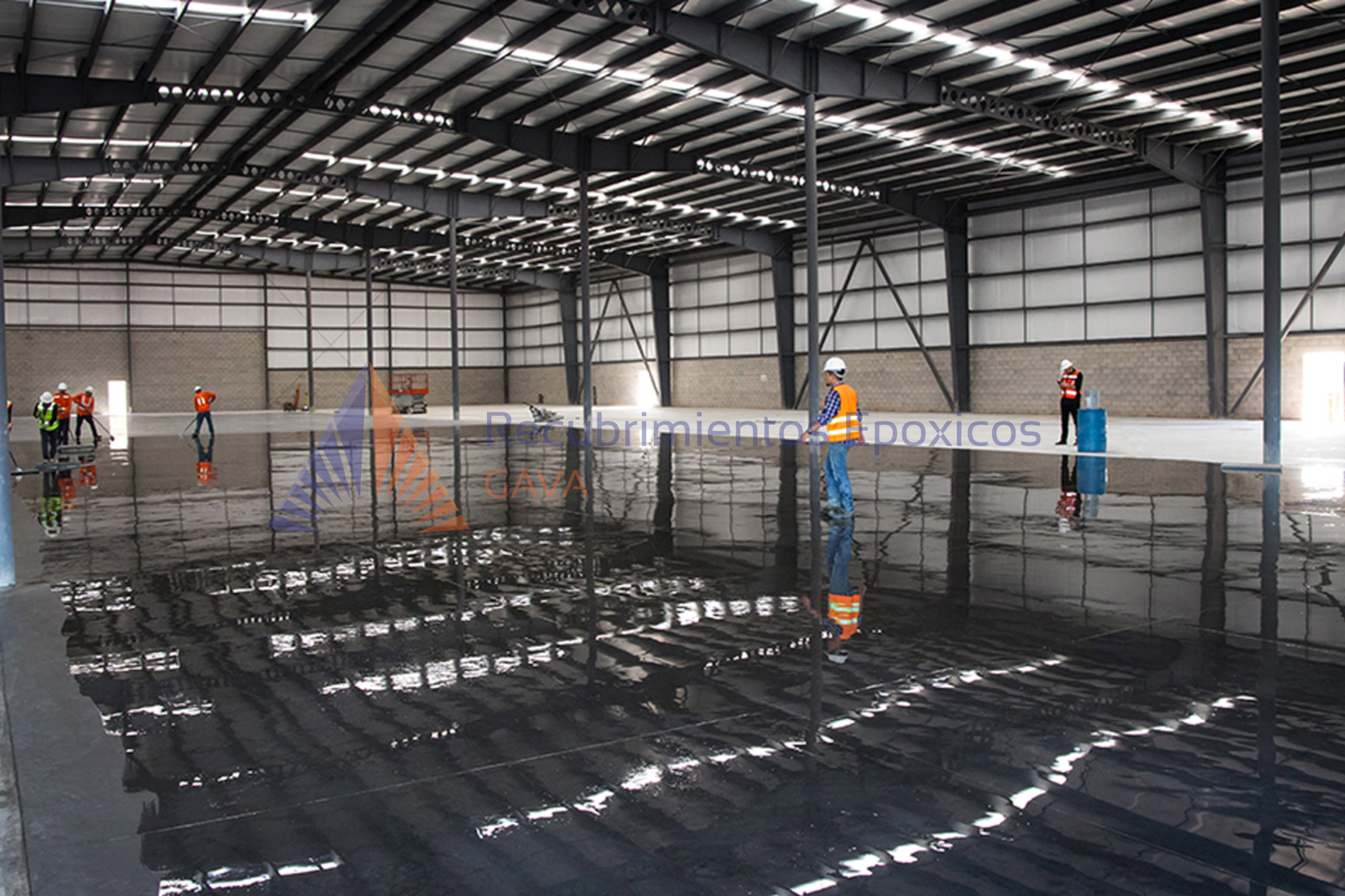 Construcción de un almacén grande con estructura de acero y paredes de paneles metálicos, empleados con cascos y chalecos reflectantes, mientras trabajan en el piso que está cubierto por agua y reflejando la estructura del techo.