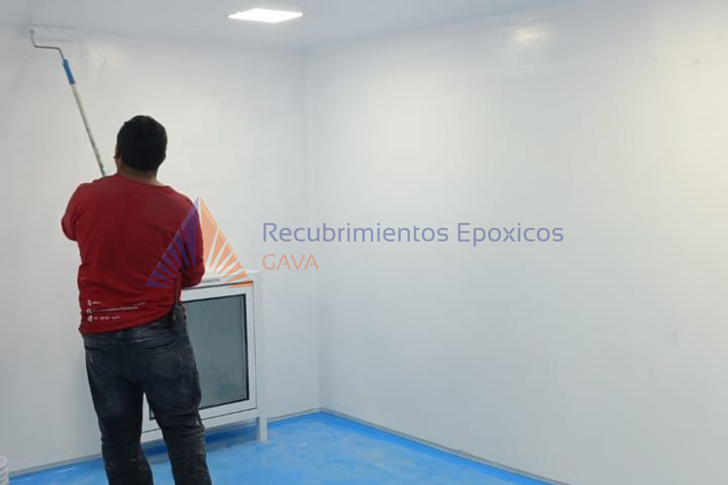 Persona pintando una pared de blanco con un rodillo, con un marco de ventana en el suelo y una pantalla en sus manos, en una habitación con suelo azul.