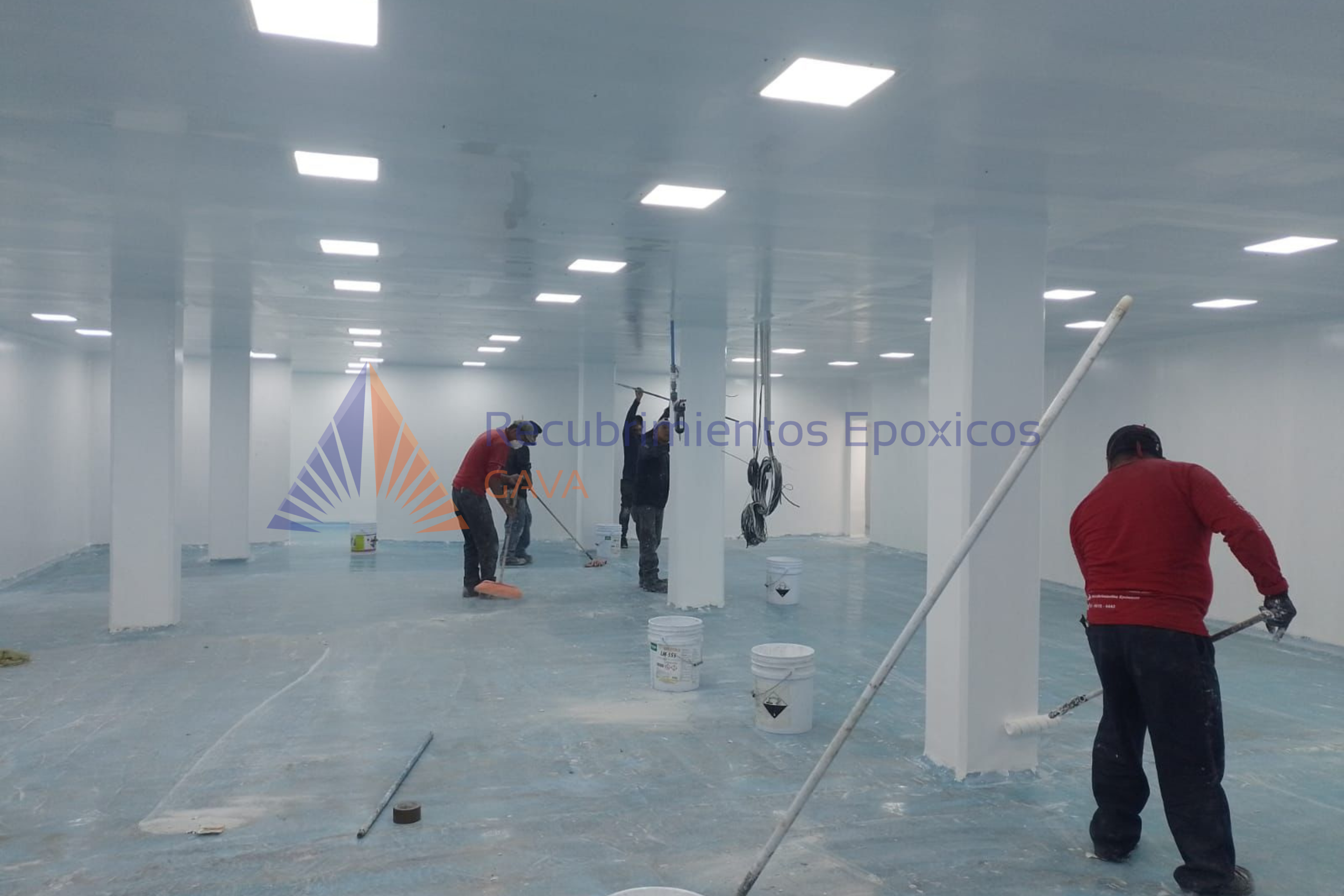 Personas trabajando en el pintado de un espacio interior con pintura epóxica blanca, con mesas, cubetas y equipo de protección en un ambiente limpio y luminoso.