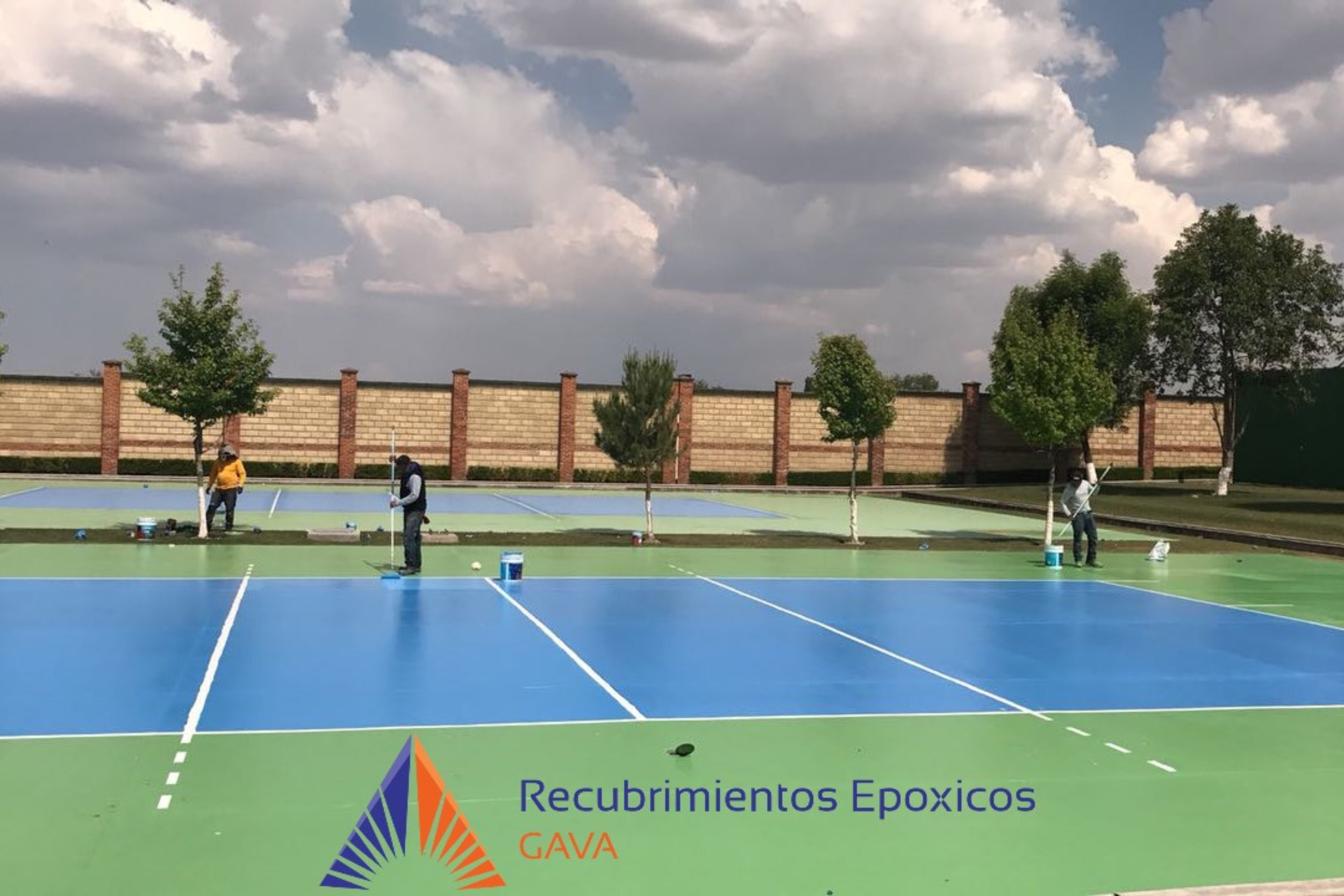 Personas pintando una cancha de tenis con recubrimientos epóxicos de colores azul y verde, en un área abierta con muro de ladrillos y árboles, bajo cielo nublado.
