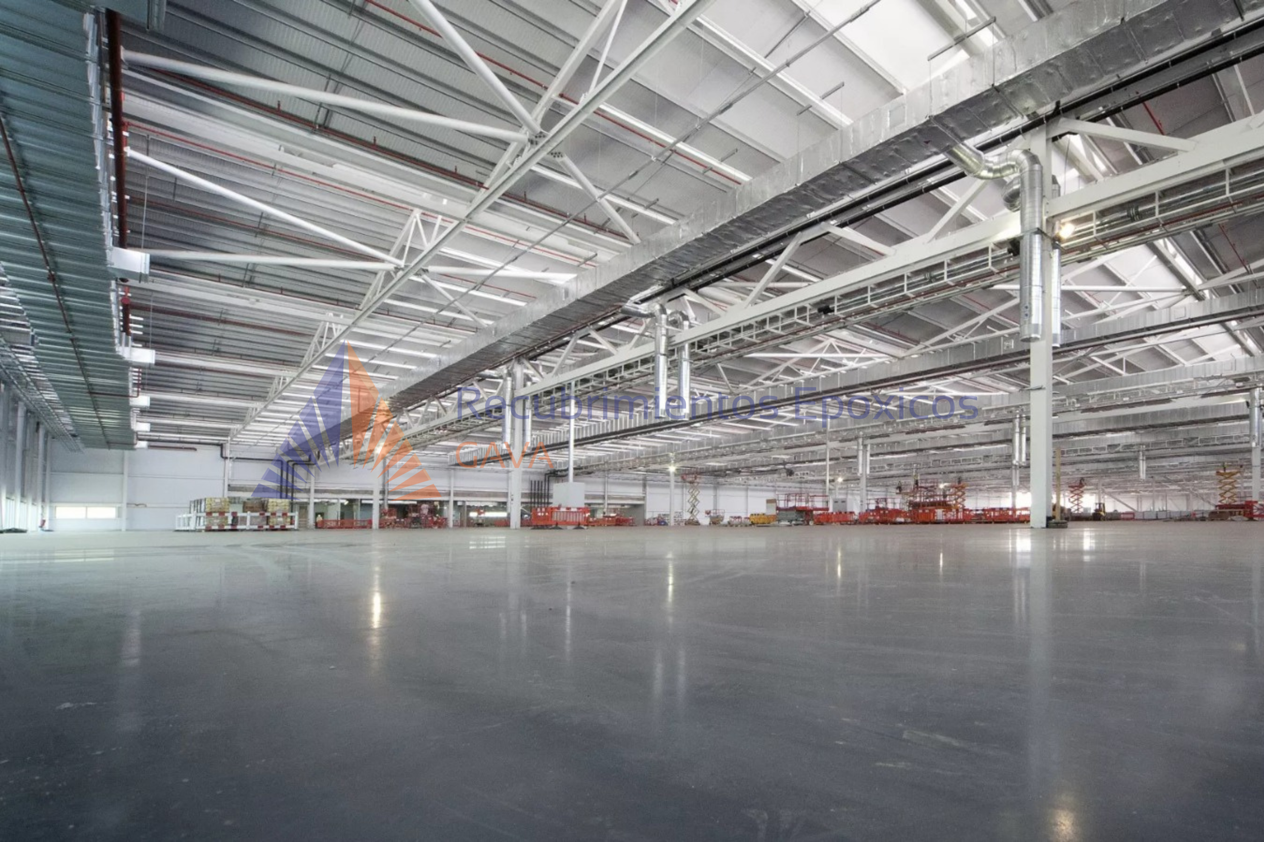 Vista de un interior de un almacén o hangar grande con estructura metálica, techos altos y amplios espacios despejados. No hay personas visibles, y en el fondo hay algunos equipos y estanterías.