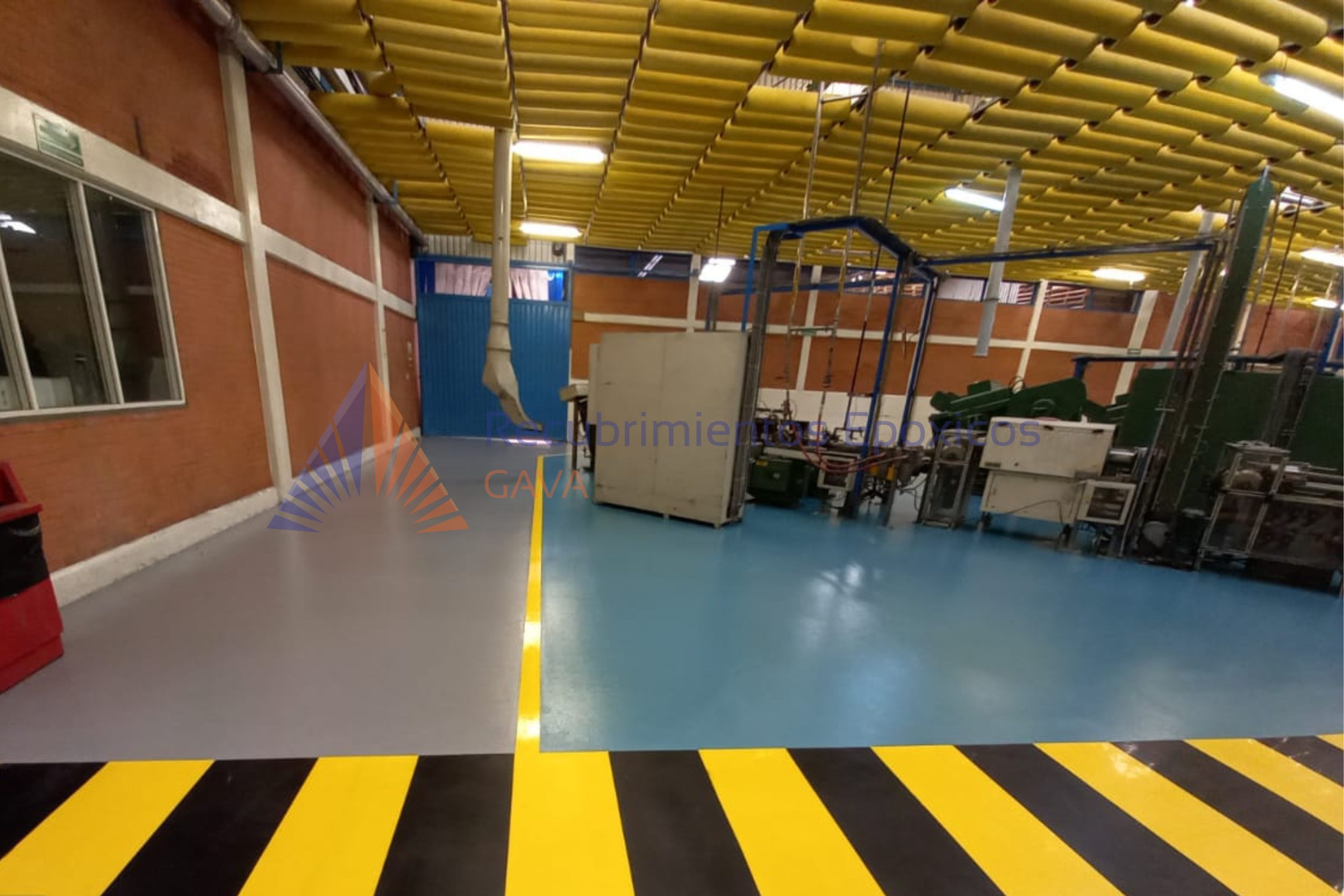 Interior de una planta industrial con techos amarillos, paredes de ladrillo y maquinaria. El suelo tiene marcas amarillas y negras. Hay ventanas y una puerta azul.