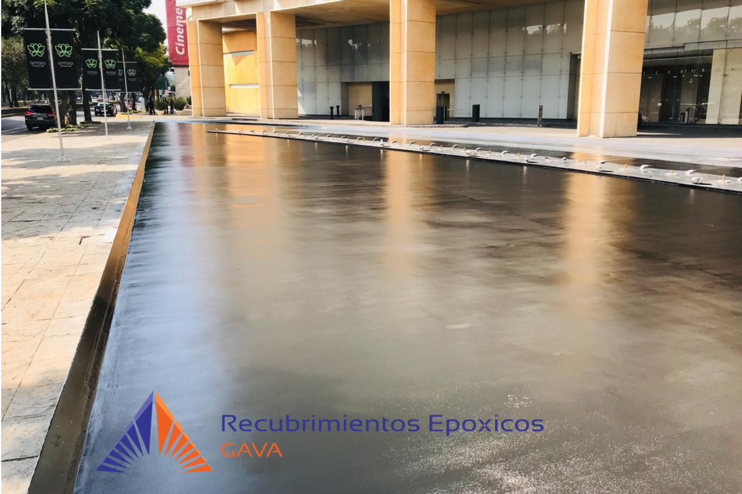 Imagen de una acera con recubrimiento epóxico en frente de un edificio moderno. Tiene un logotipo y texto que dice 'Recubrimientos Epóxicos GAVA' en la parte inferior izquierda.