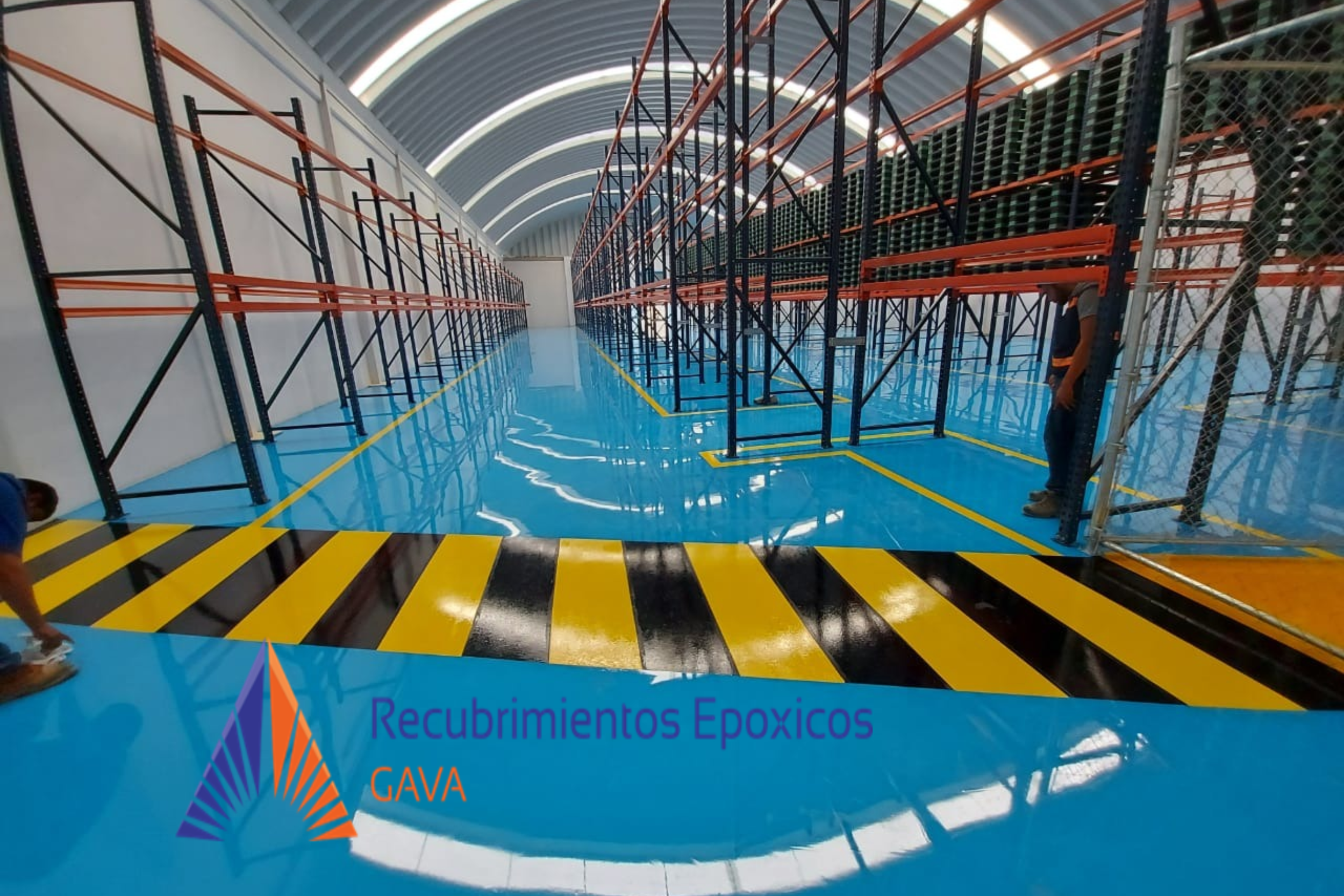 Interior de una estructura de almacenamiento con estanterías metálicas y pisos de color azul, señalización de precaución en amarillo y negro, y un cartel que dice "Recubrimientos Epóxicos GAVA".