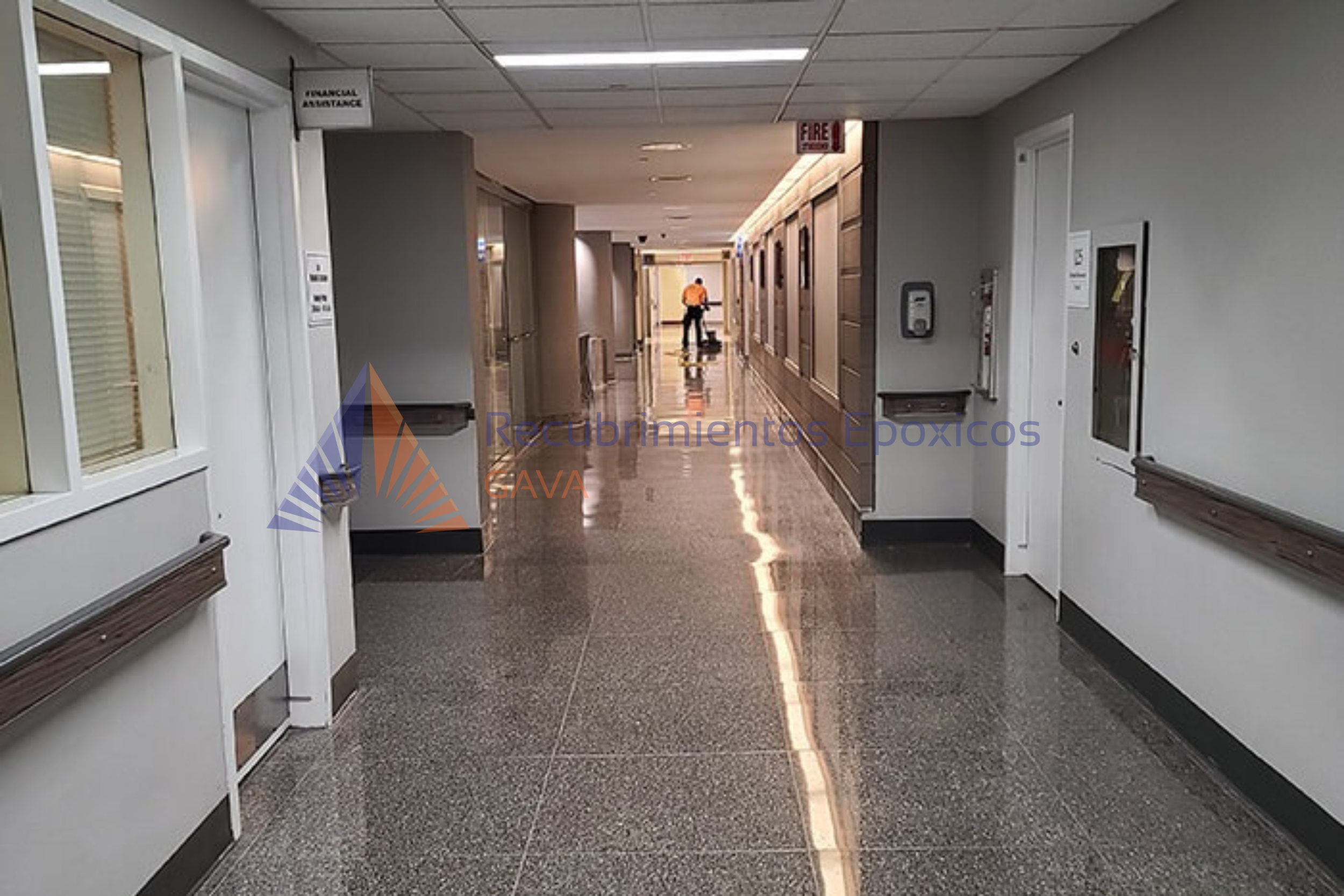 Pasillo en edificio con paredes grises, pisos de granito, señales de emergencia y una persona con chaqueta naranja y maletín en el fondo.