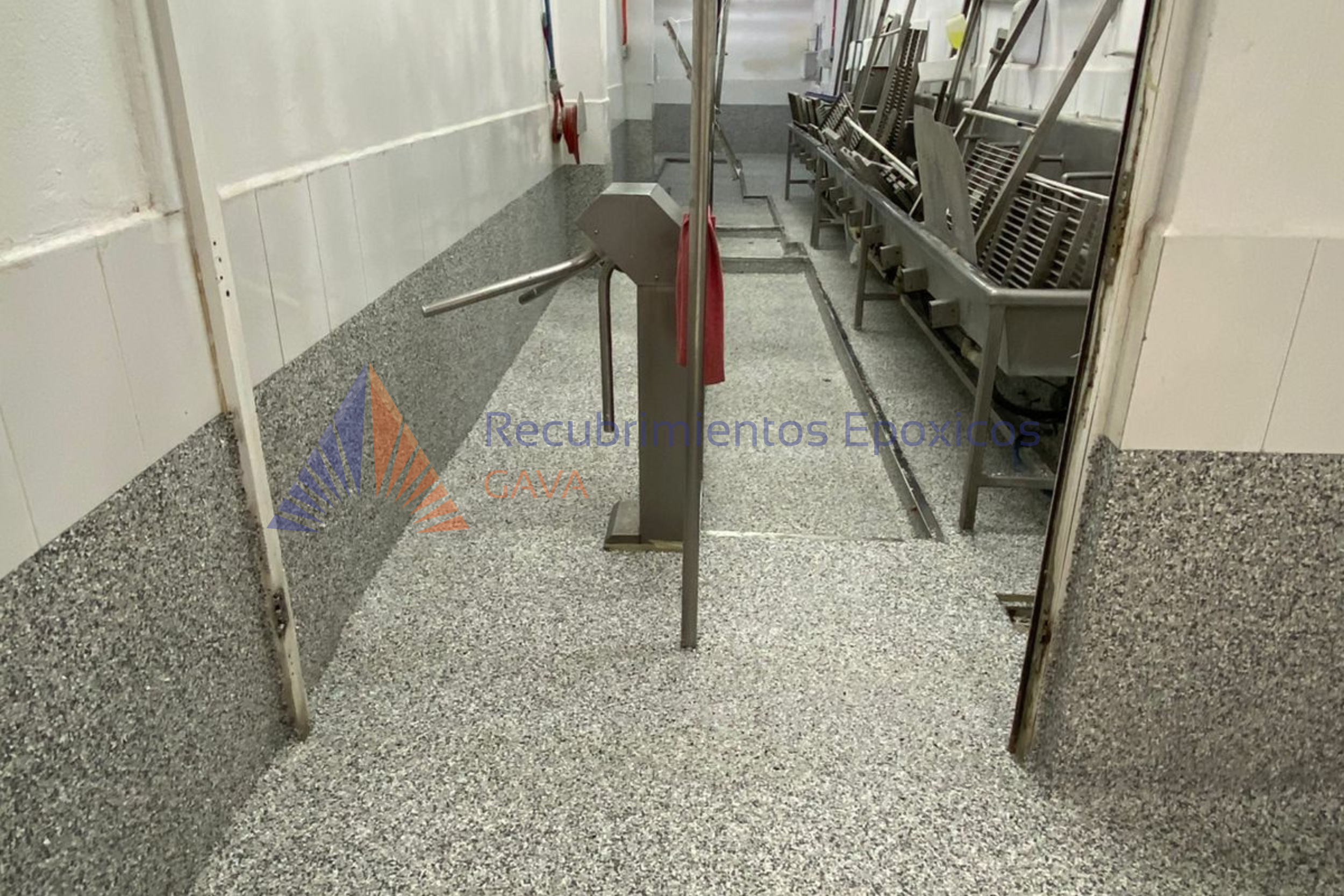 Puerta abierta a un pasillo de un edificio con mesas metálicas apiladas del lado derecho y una barrera de control en el centro de la imagen.