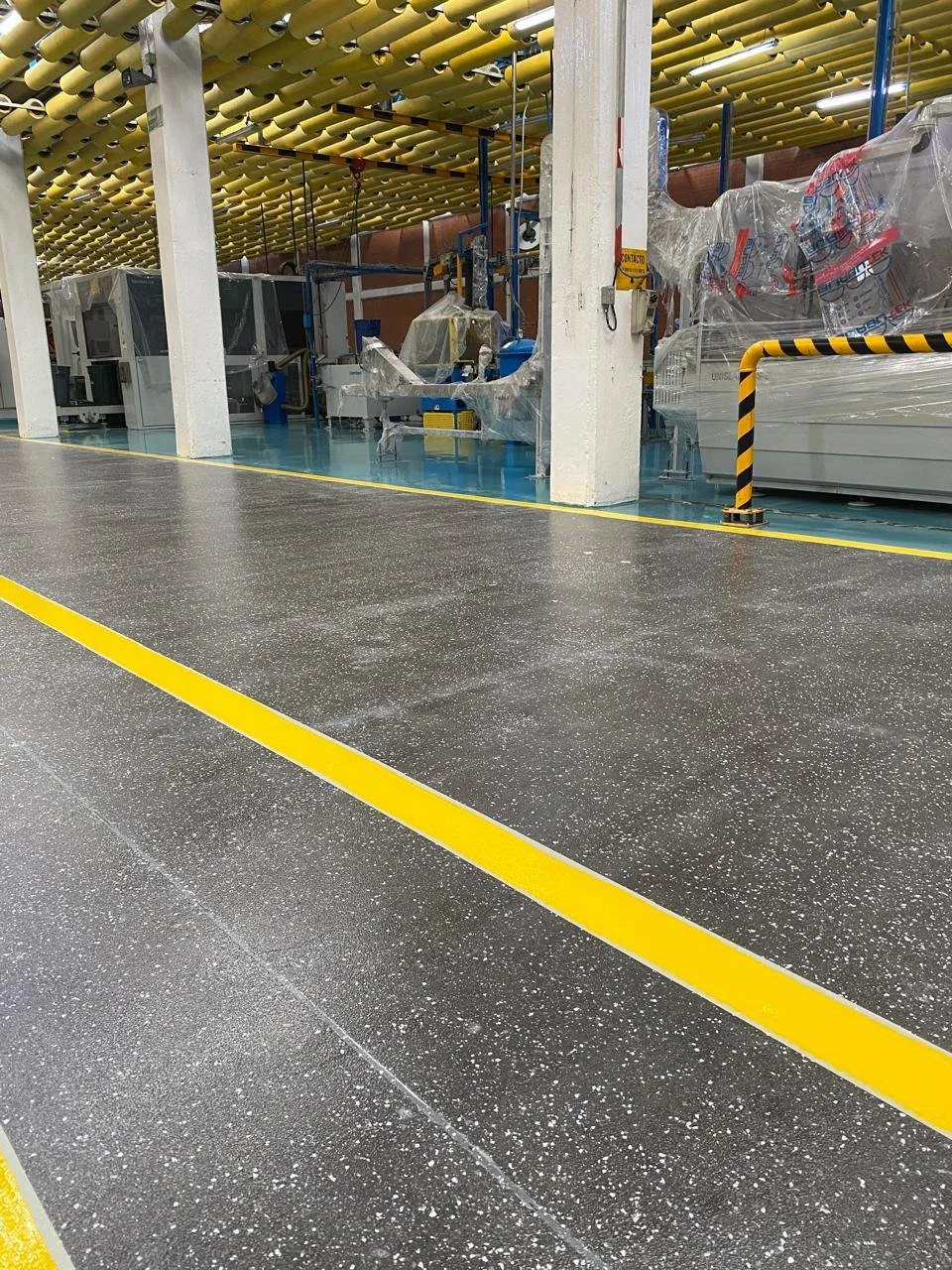 Interior de una fábrica con maquinaria cubierta parcialmente con plástico, piso negro con puntos blancos y líneas amarillas.