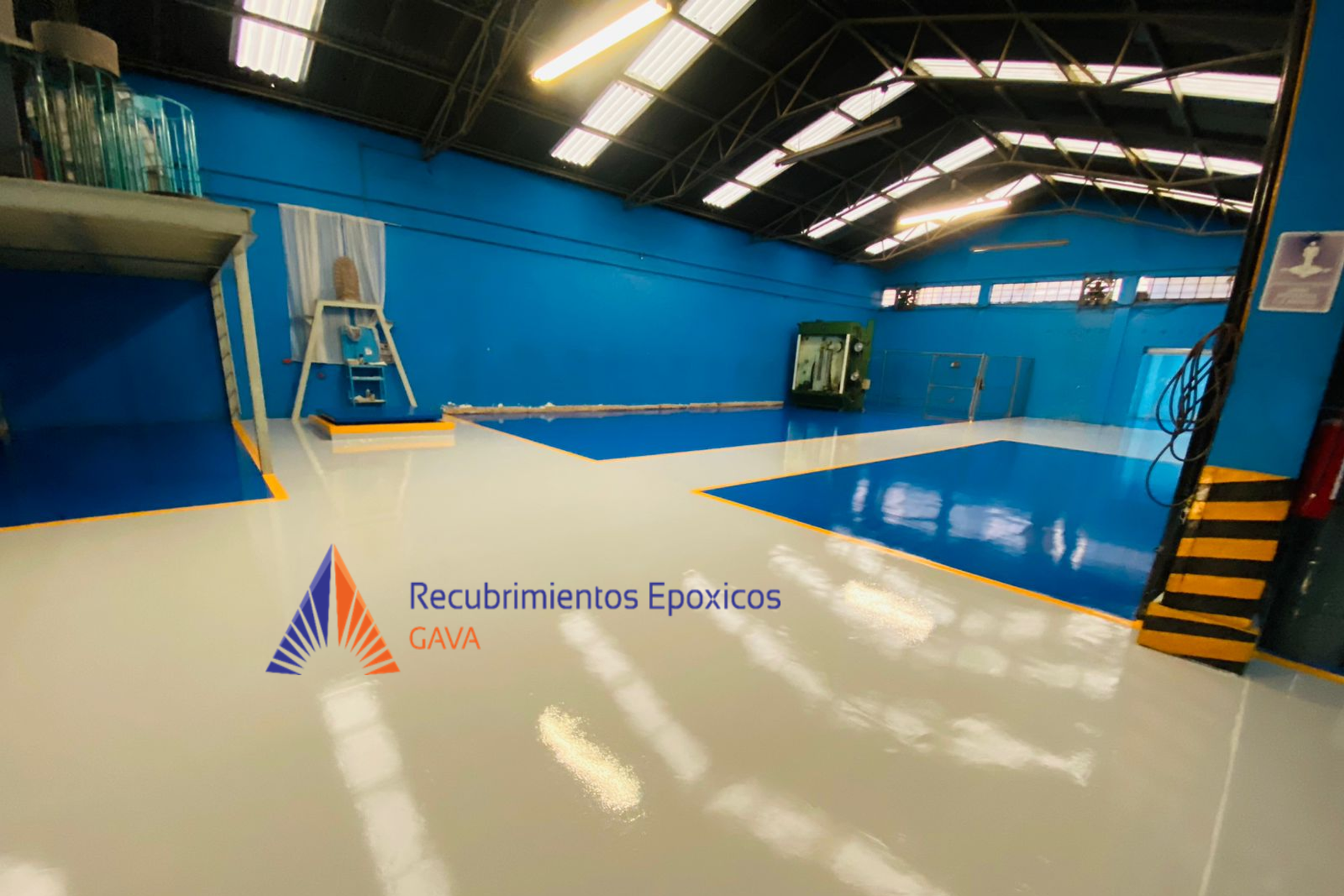 Un espacio industrial con paredes y techo de color azul, suelo brillante de color beige con marcas negras y anaranjadas, y maquinaria en el fondo. En la parte inferior izquierda, logotipo que dice "Recubrimientos Epóxicos GAVA" con un diseño colorido