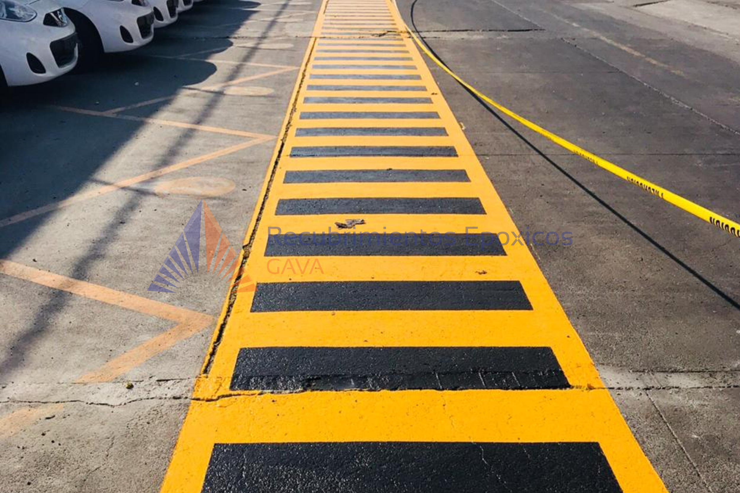 Línea amarilla y negra en el suelo que indica una zona de advertencia en un estacionamiento con coches blancos alineados a la izquierda.