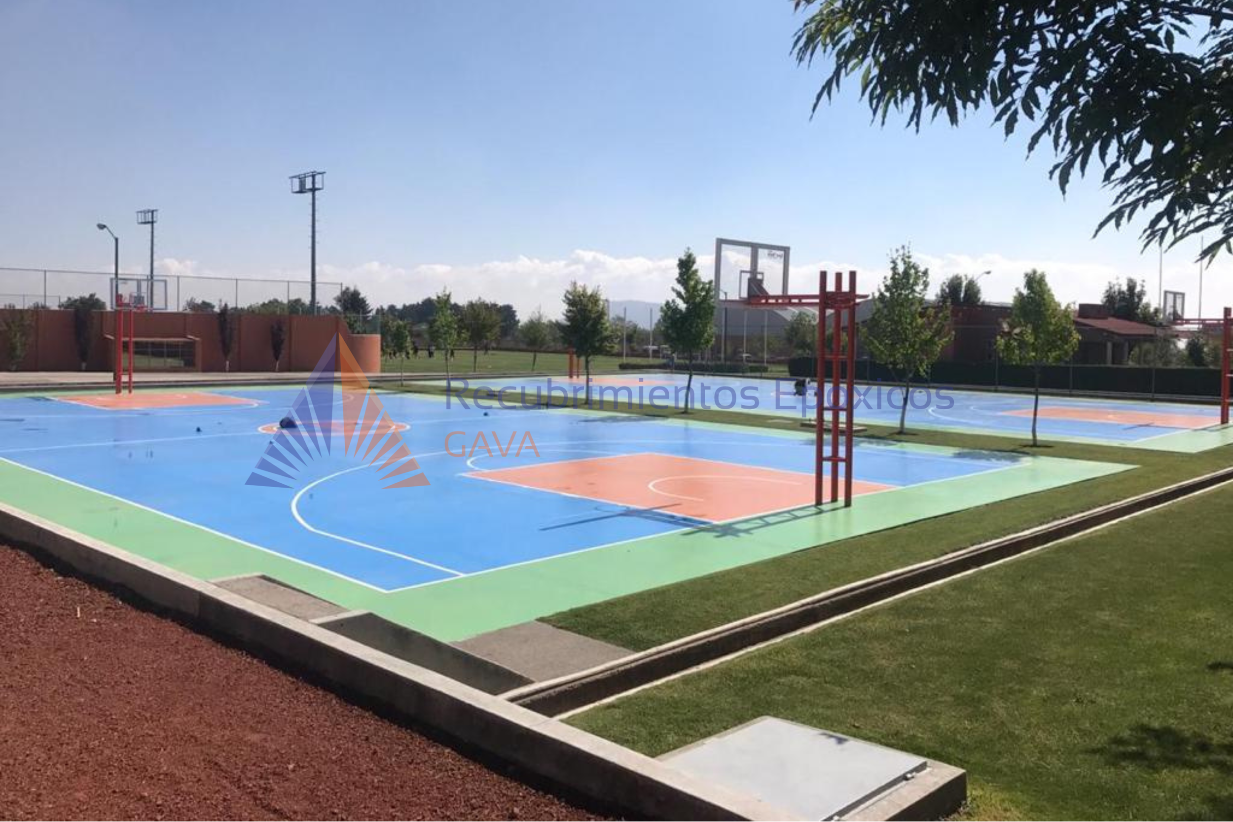 Cancha de baloncesto exterior con superficies de colores azul y naranja, rodeada de árboles verdes y césped, en un día soleado