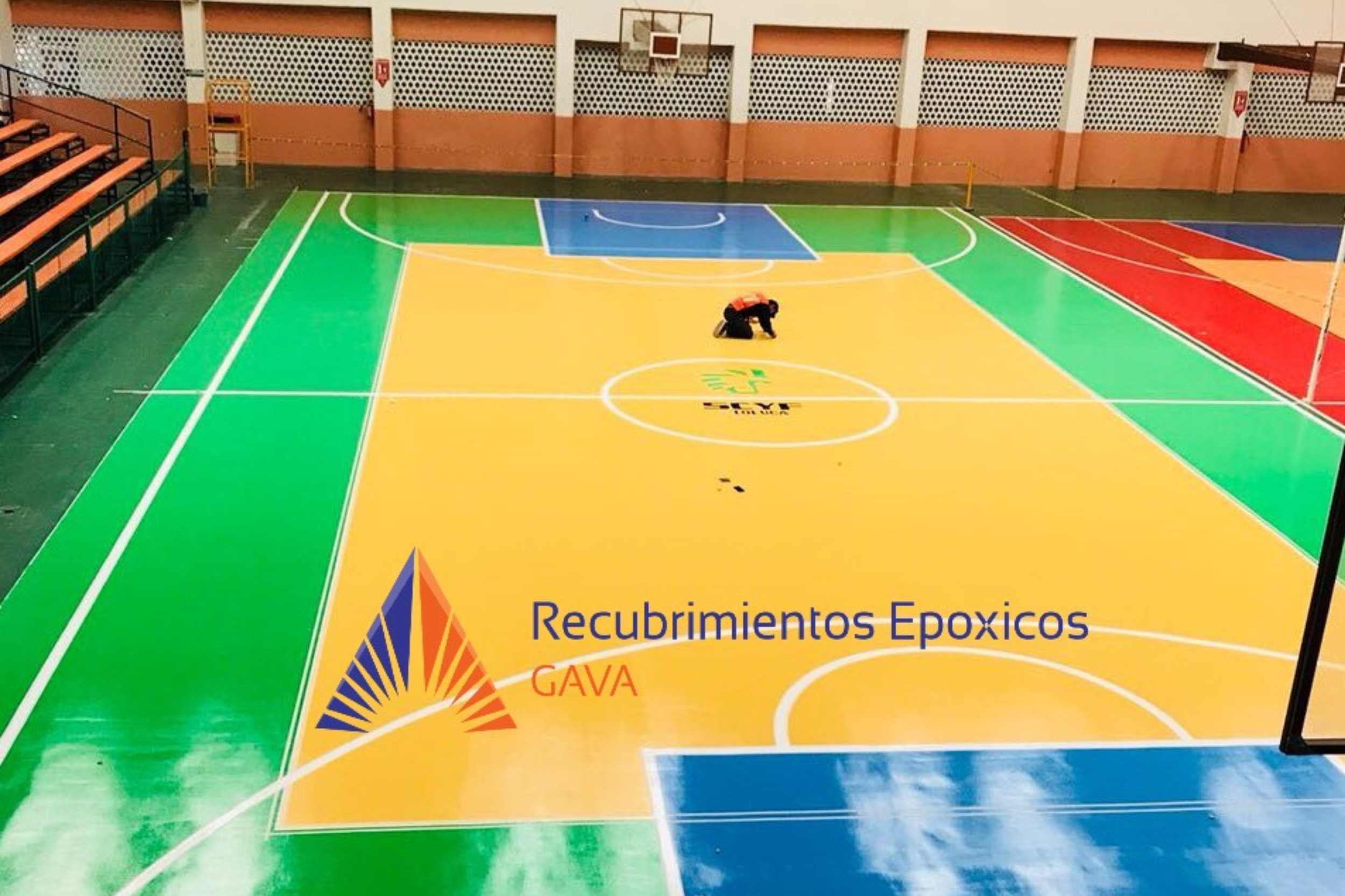 Vista de una cancha de baloncesto en una sala cubierta, con áreas de diferentes colores (azul, verde, amarillo, rojo). En el centro, hay una persona arrodillada limpiando o pintando el suelo, con la marca y logo de la cancha visible y el texto "Regra