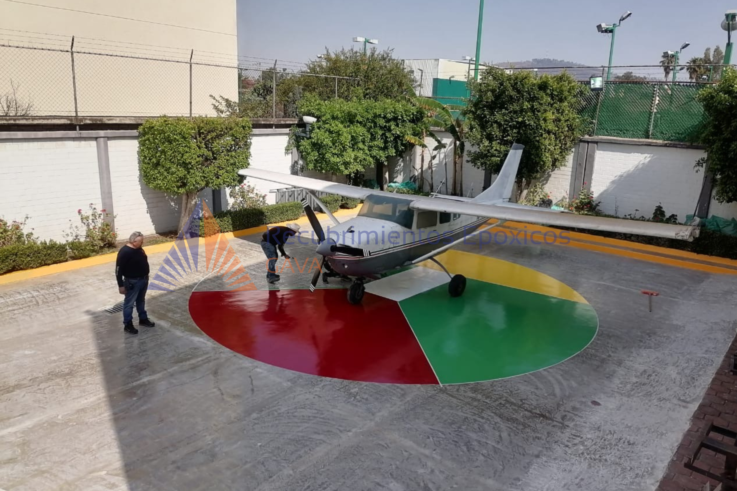Helicóptero con colores de la bandera de México, en un helipuerto rodeado de árboles y paredes blancas, con dos personas observando.