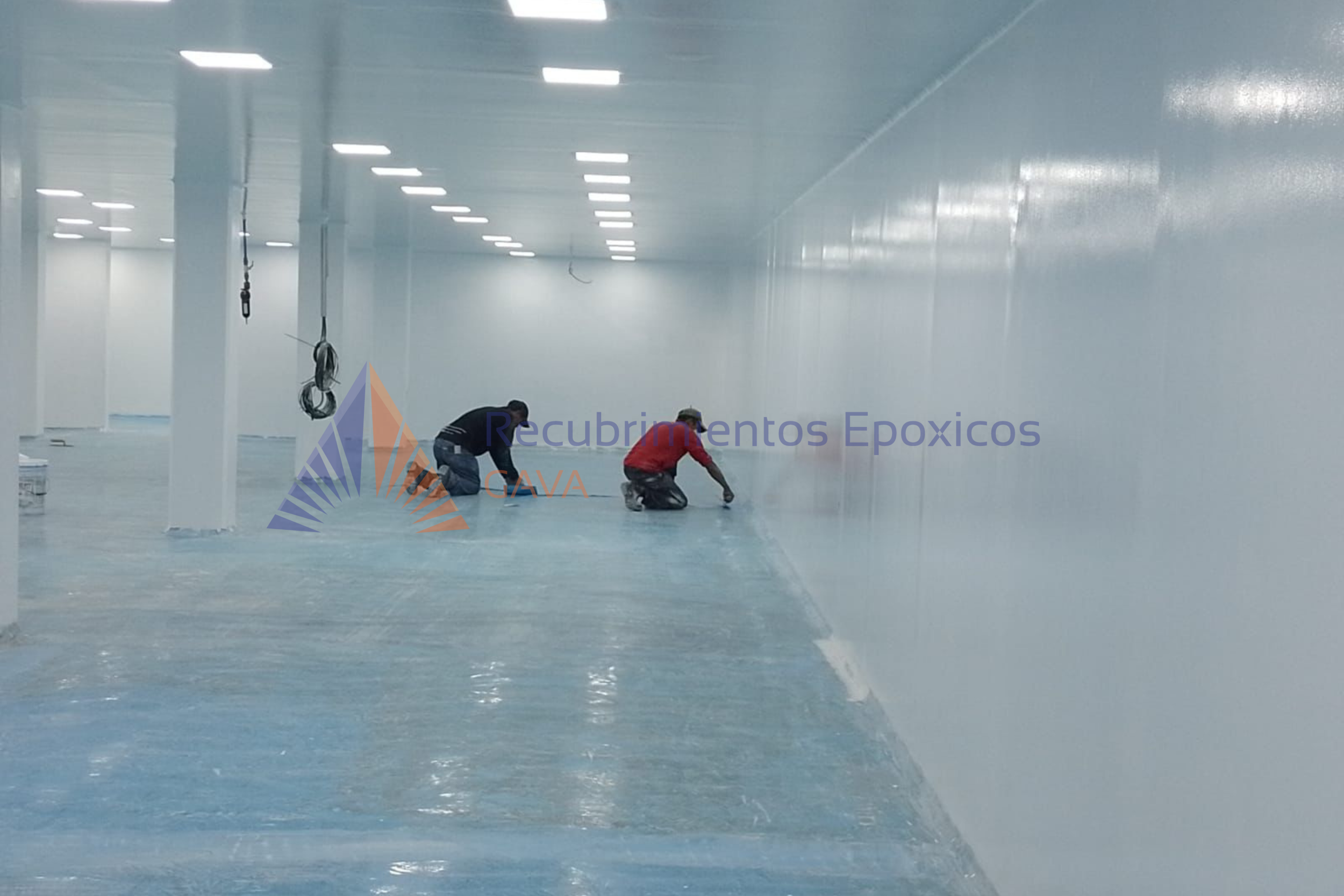 Dos trabajadores aplicando recubrimiento epóxico en el suelo de un espacio interior grande, con paredes y techo blancos y luces LED en el techo.