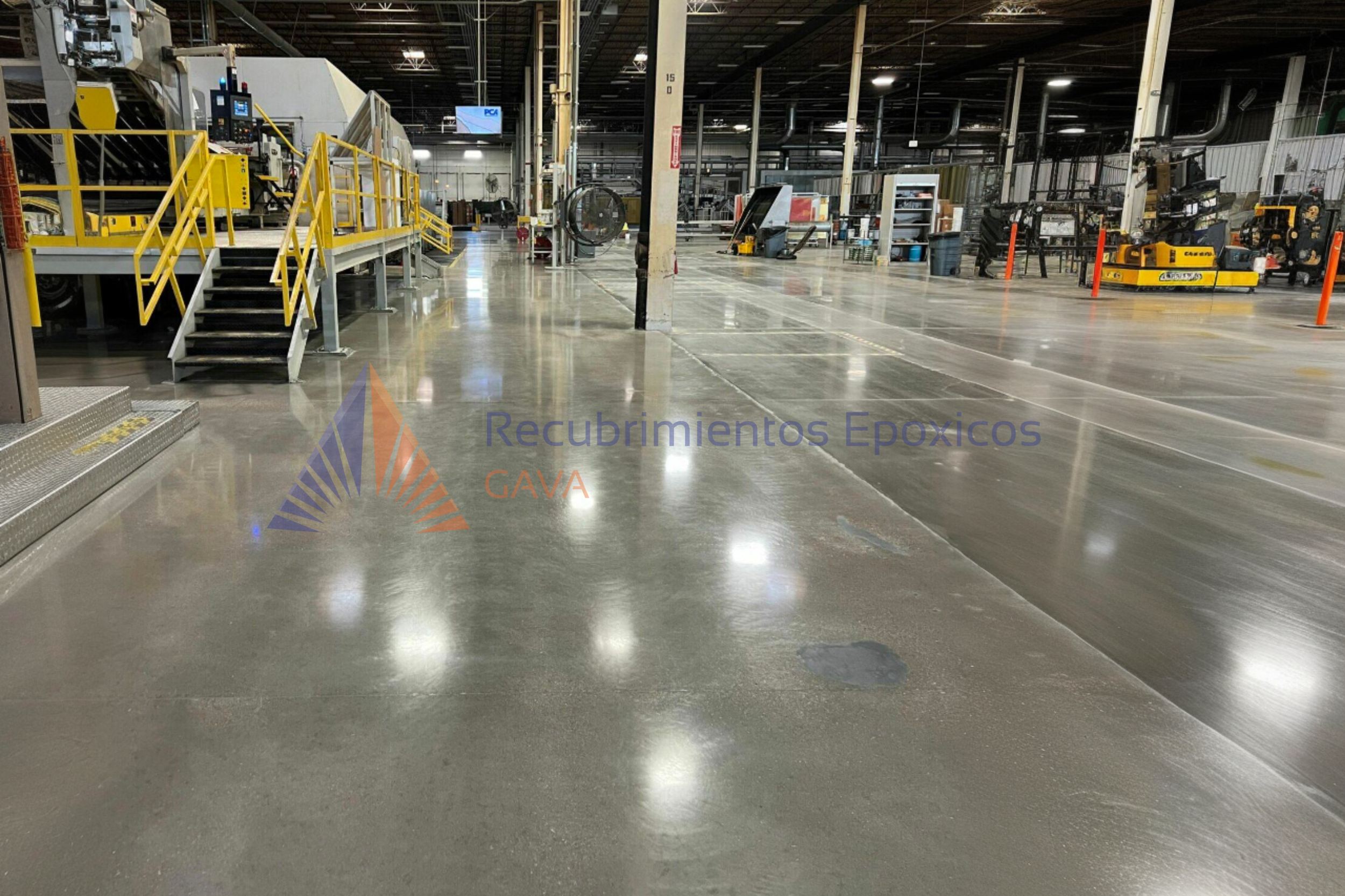 Amplio almacén industrial con maquinaria y equipos en proceso, pisos de concreto pulido y señalización de seguridad, con el logo de Recubrimientos Epóxicos GAVA en la esquina inferior izquierda.