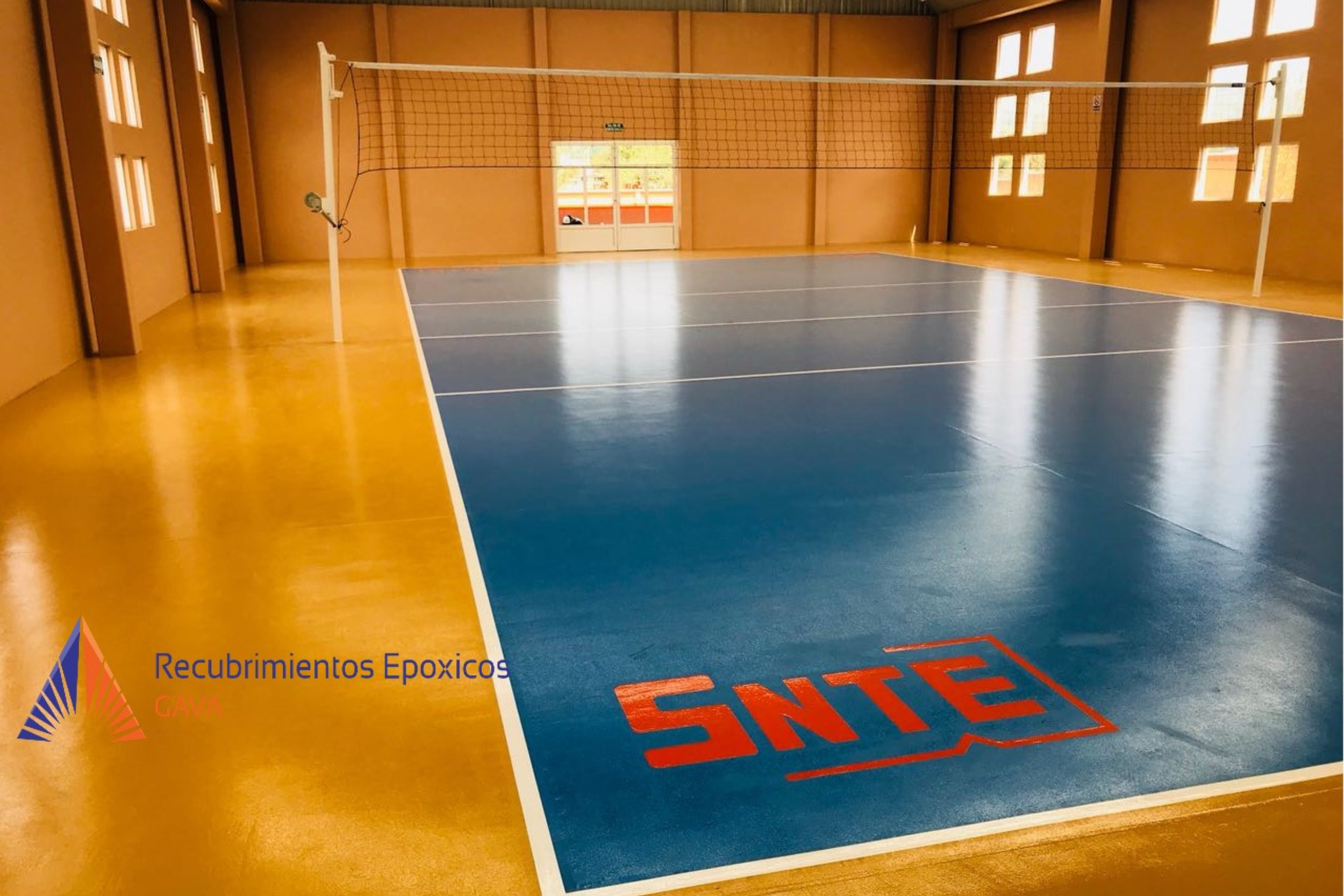 Pista de voleibol en una cancha cubierta con suelo de color amarillo y línea blanca, red de voleibol en el centro y marca de advertencia en la esquina inferior derecha que dice 'SINE'.