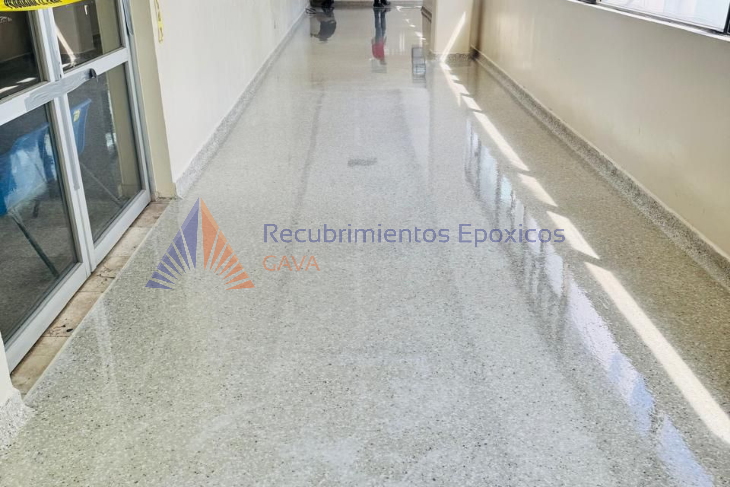 Pasillo interior con piso de recubrimiento epóxico color gris claro y paredes blancas, con ventanas y puerta de vidrio a la izquierda y personas caminando al fondo.