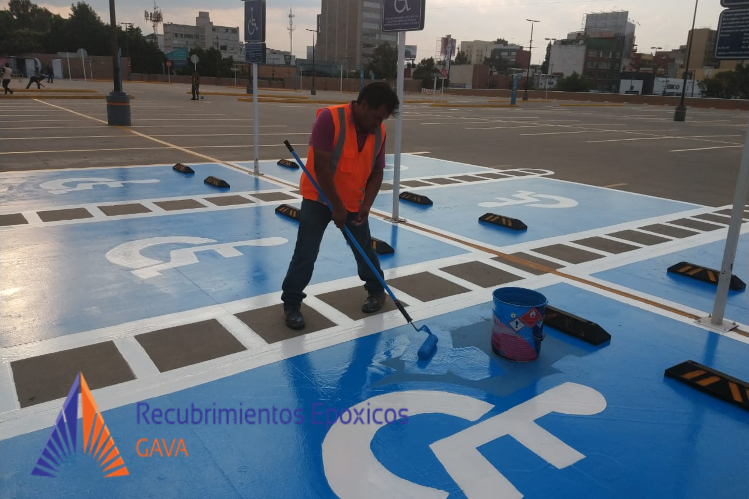 Una persona pintando un espacio de estacionamiento para discapacitados en un estacionamiento grande, con marcas en el suelo y señalizaciones, usando pintura azul y un rodillo.