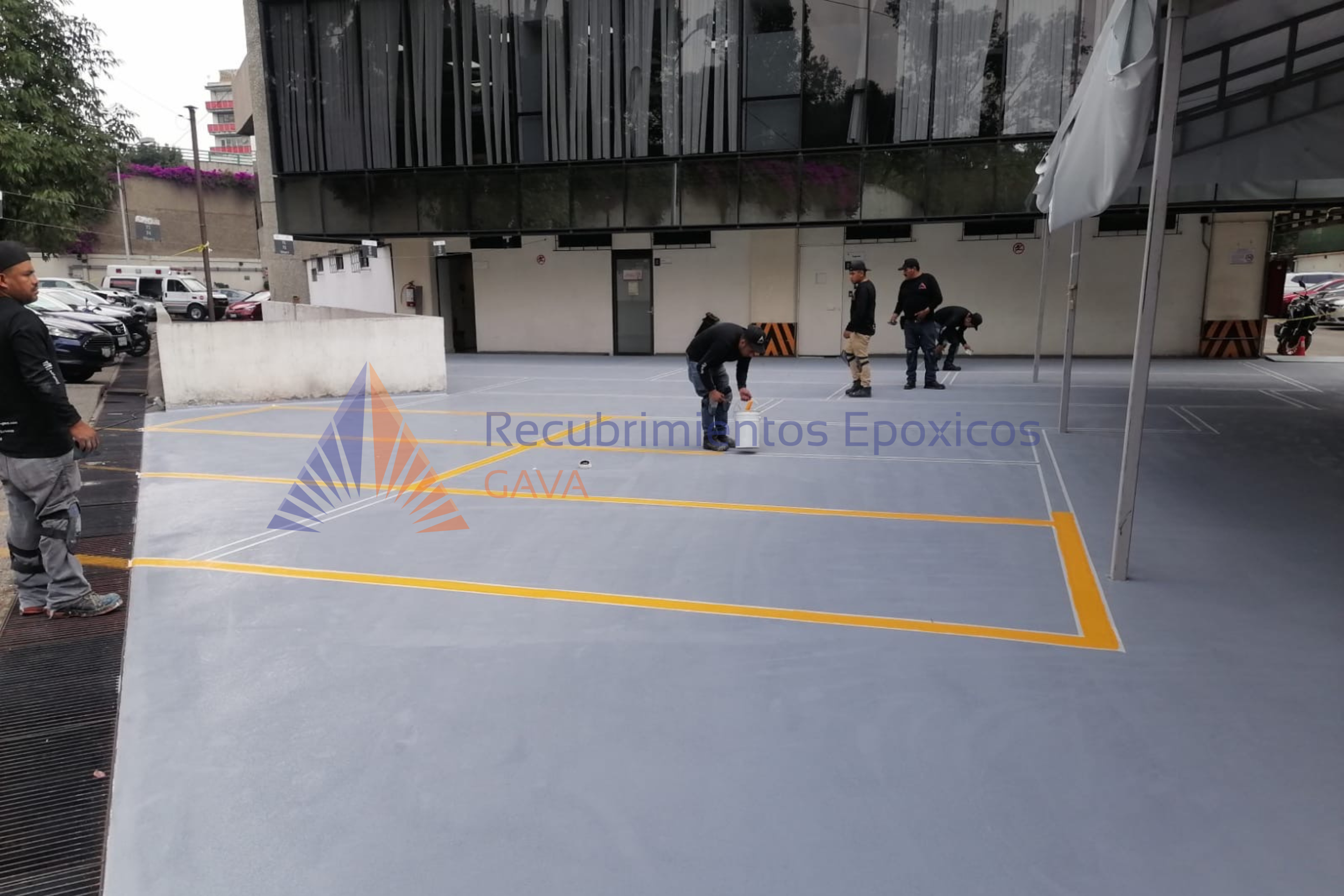 Hombres trabajando en la instalación o mantenimiento de pintura epóxica en un estacionamiento al aire libre frente a un edificio moderno.